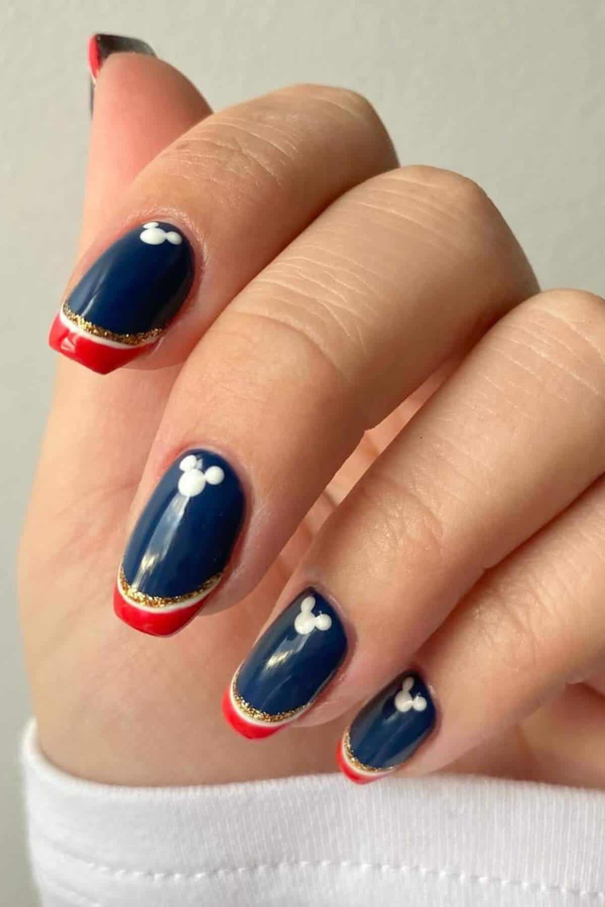 A hand with navy blue nails featuring white Mickey Mouse head shapes, red tips, and a thin gold line near the edge of each nail.