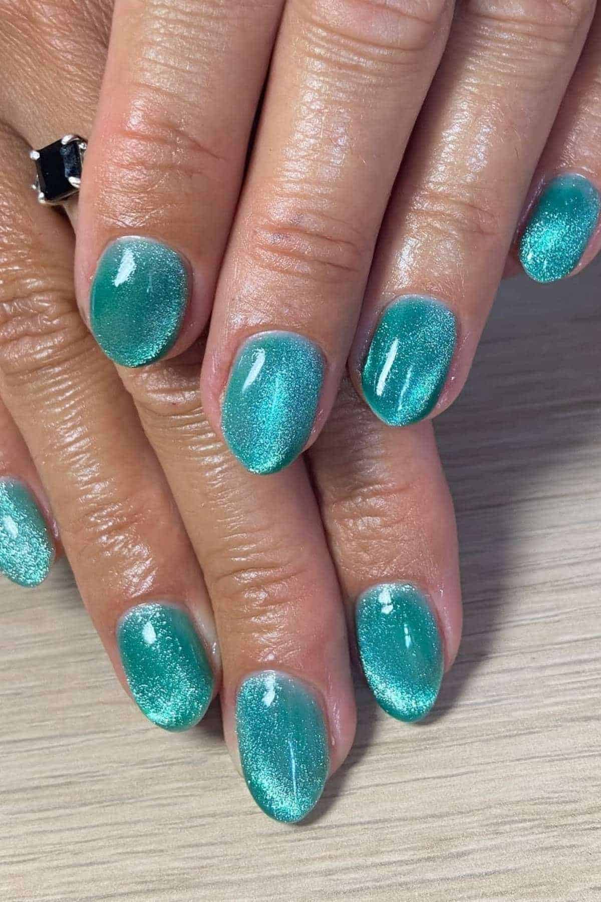 Close-up of hands with short, rounded nails painted in a shimmery teal blue polish, resting on a light-colored surface.