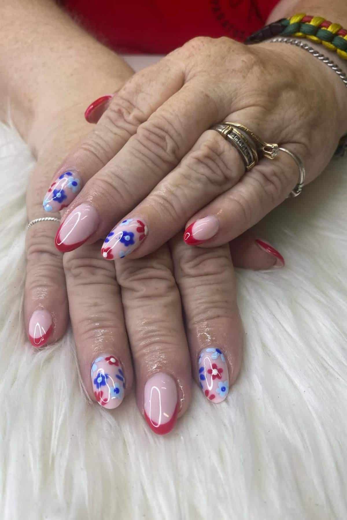 Close-up of hands with manicured nails featuring red tips and blue and red floral designs, resting on a white textured surface.