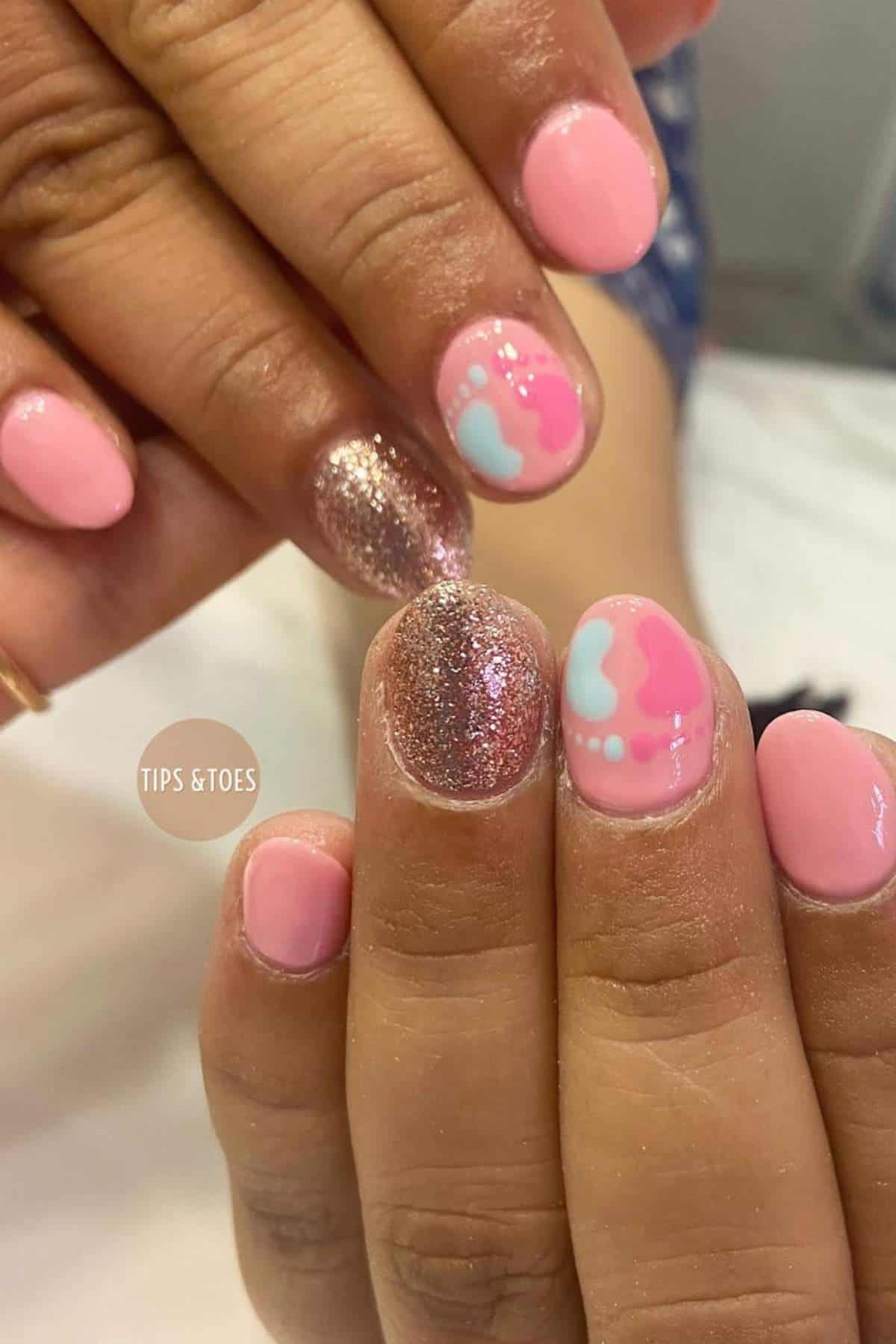 Close-up of hands with pink and glittery nail polish; some nails feature baby blue and pink footprint designs.