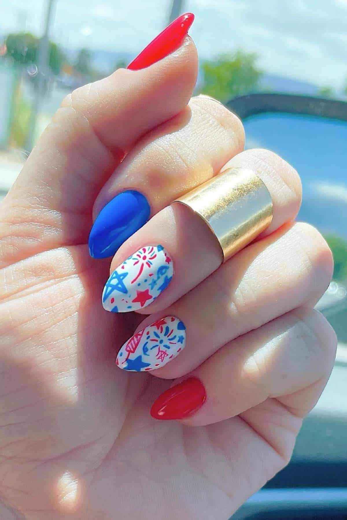A hand with manicured nails painted in red, blue, and white, featuring star and firework designs, holding up fingers with a gold ring.
