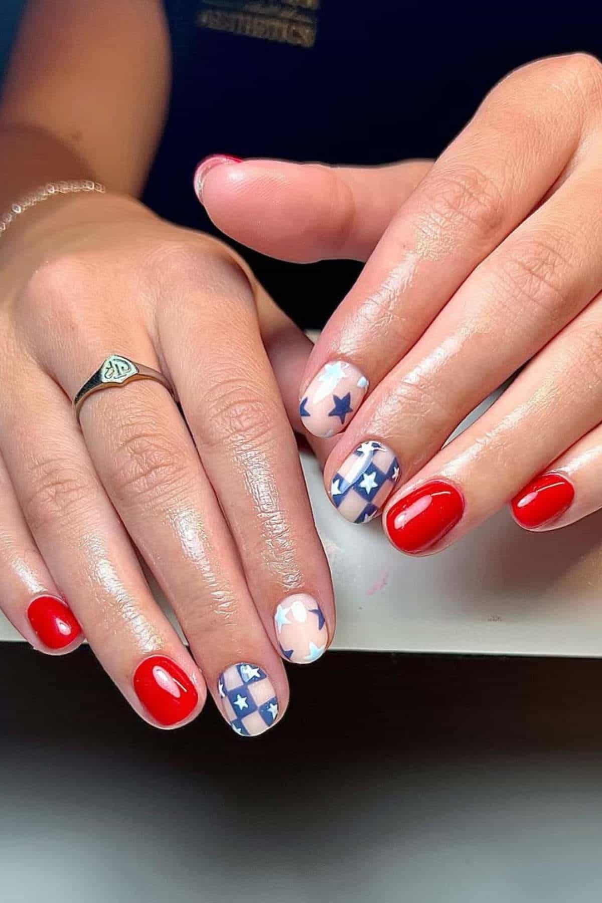 Hands with short, rounded nails painted red, with two accent nails featuring blue and white stars and stripes, inspired by the American flag.