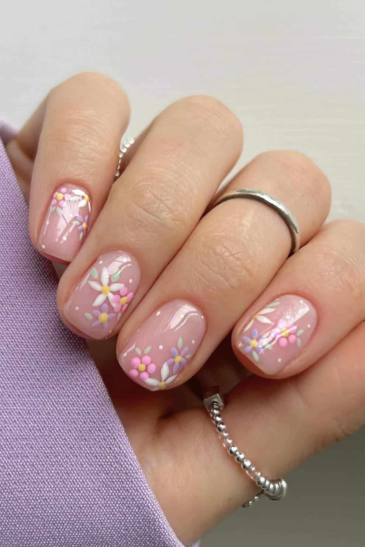 A hand with short, glossy pink nails featuring small white, yellow, and pink flower designs. The person is wearing silver rings and a lavender-colored sleeve.
