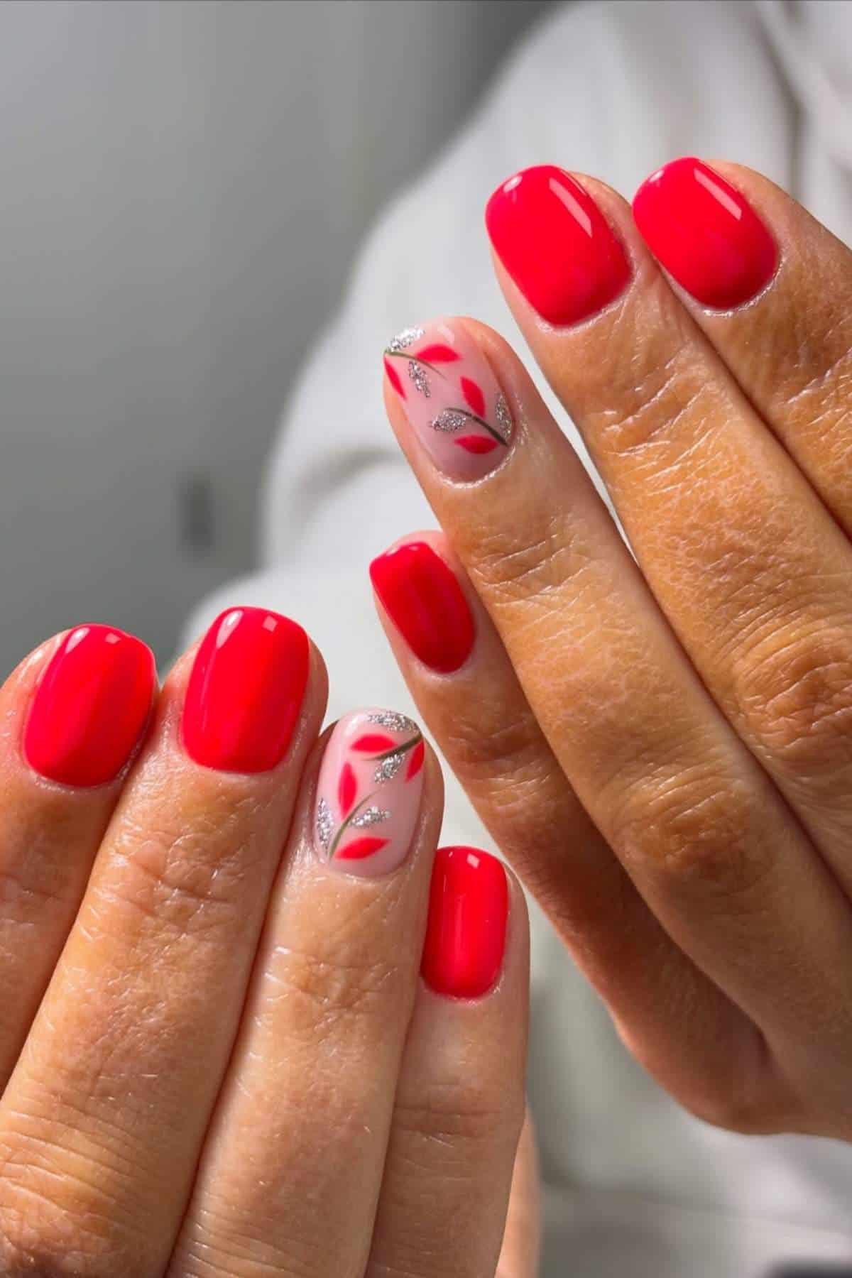 Hands with short, glossy red nails; two accent nails feature a nude base with silver and red leaf designs.
