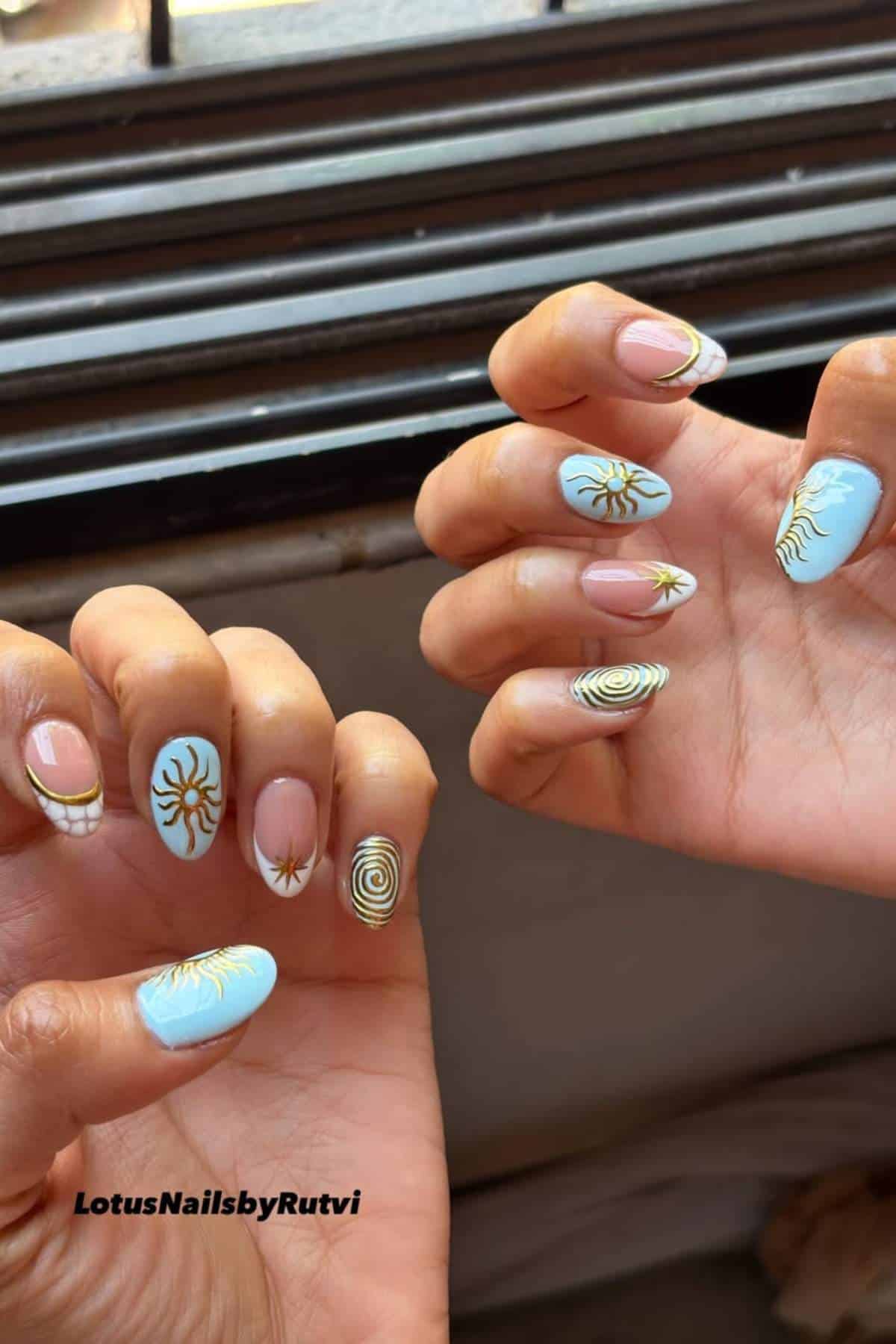 Hands with almond-shaped nails featuring blue and nude bases, decorated with gold sun, star, and spiral designs. Nails by LotusNailsbyRutvi.
