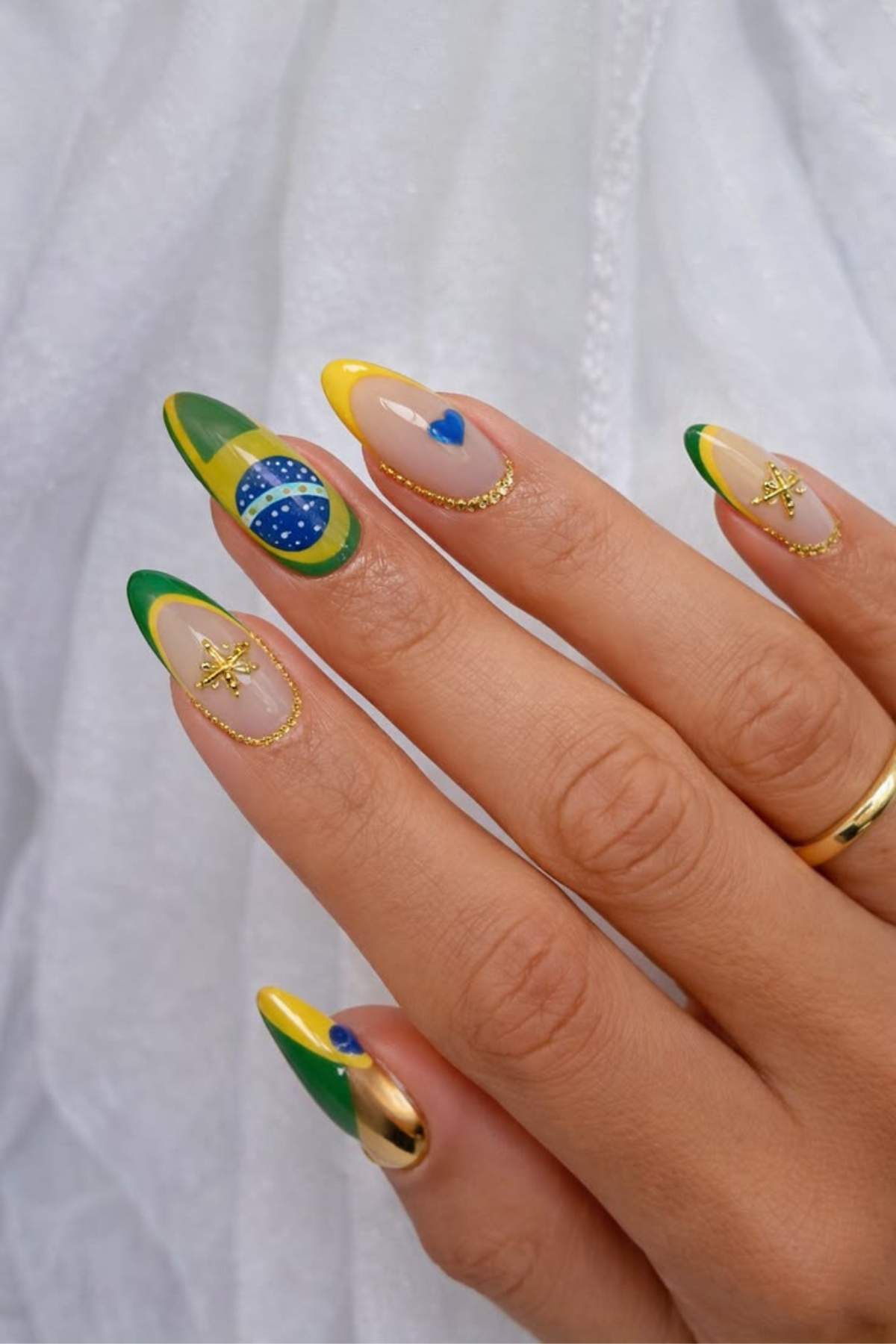 A hand with long, almond-shaped nails painted in green, yellow, and blue designs inspired by the Brazilian flag, featuring gold and heart accents.