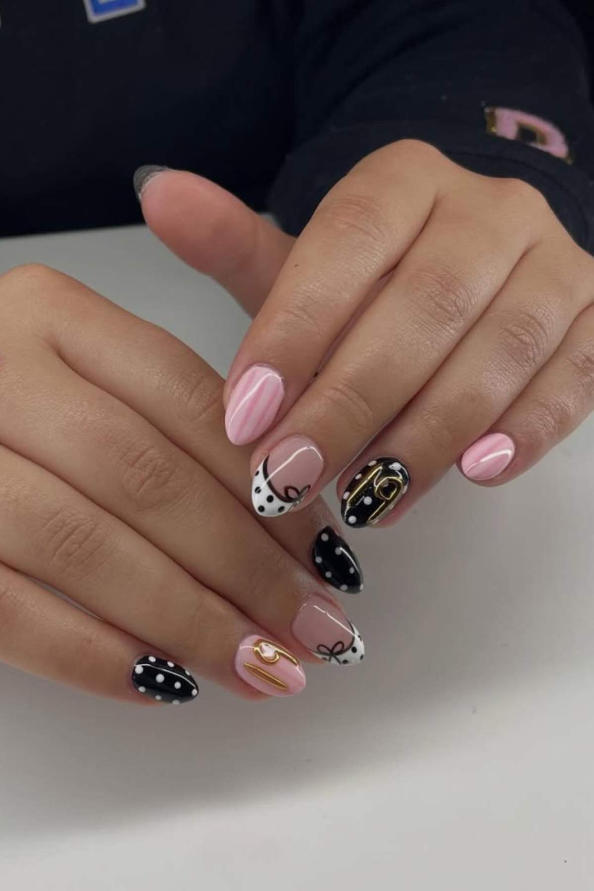 Hands with manicured nails featuring a mix of pink, black, and white designs, including polka dots, stripes, abstract lines, and gold accents.