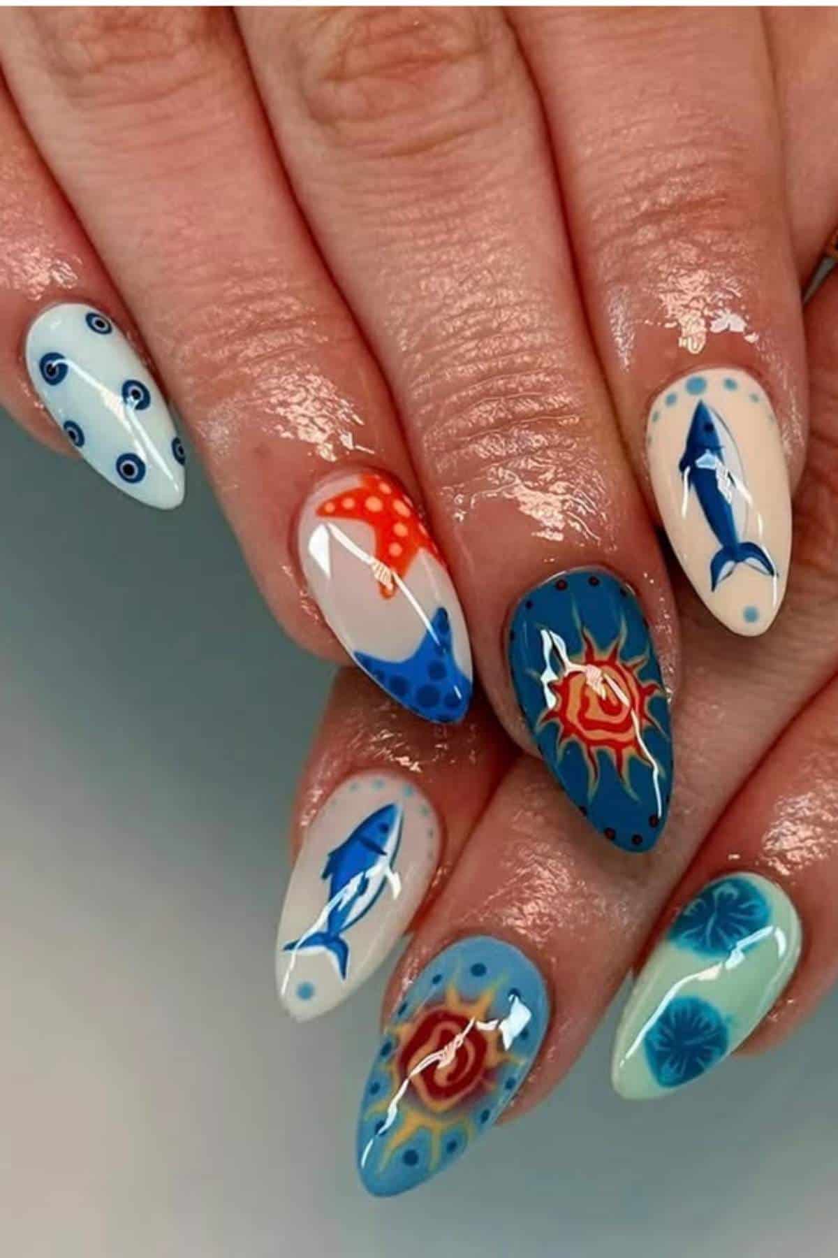 A hand with long, almond-shaped nails featuring ocean-themed nail art, including sharks, waves, fish, and abstract underwater designs in blue, white, red, and orange.