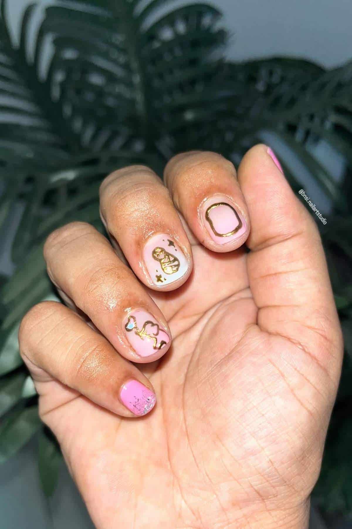 A hand with short nails painted in pink and white polish, featuring gold abstract designs and accents, held against a leafy background.