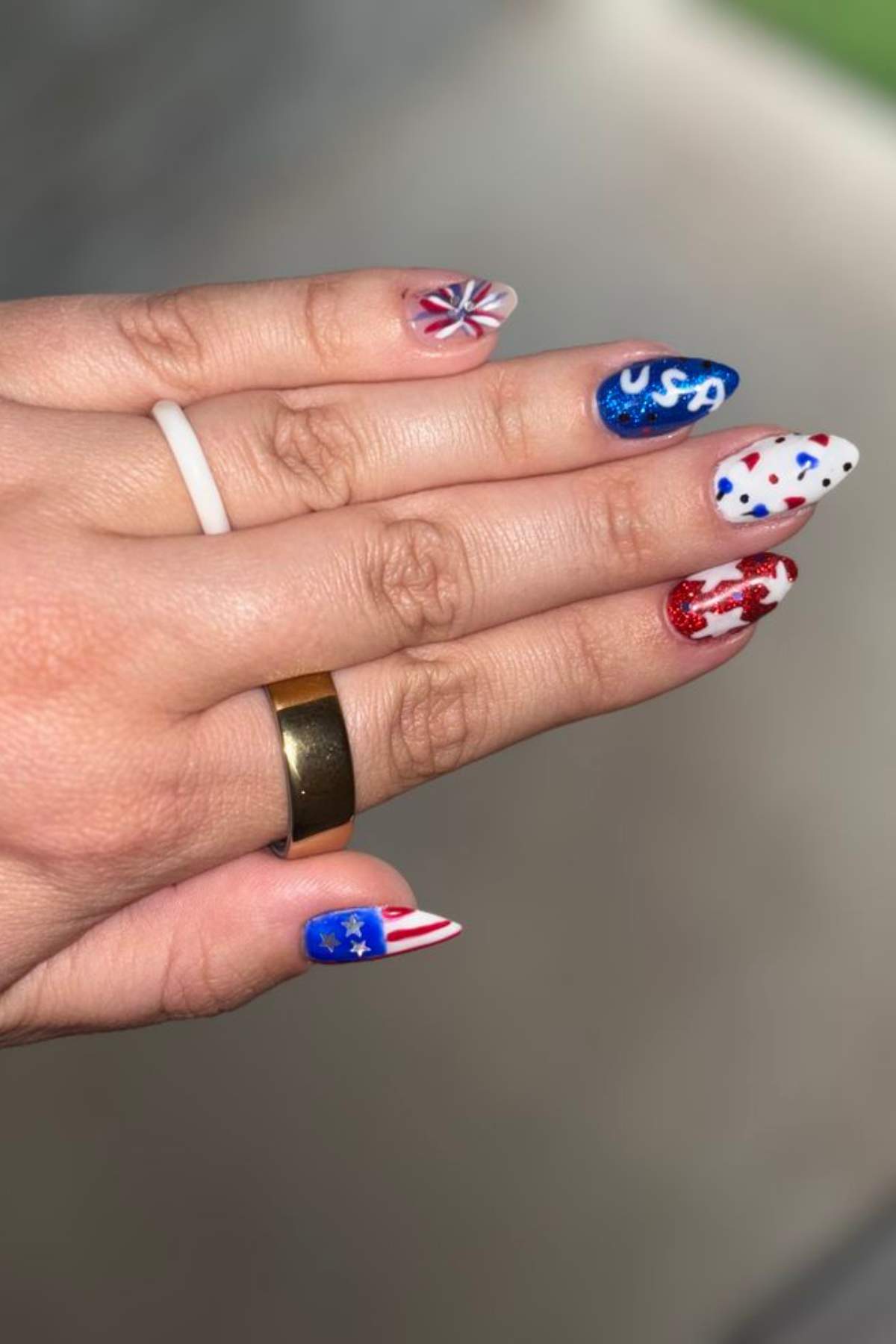 A hand with manicured nails painted in red, white, and blue patriotic designs, including stars, stripes, and "USA" text. Two rings are on the fingers.