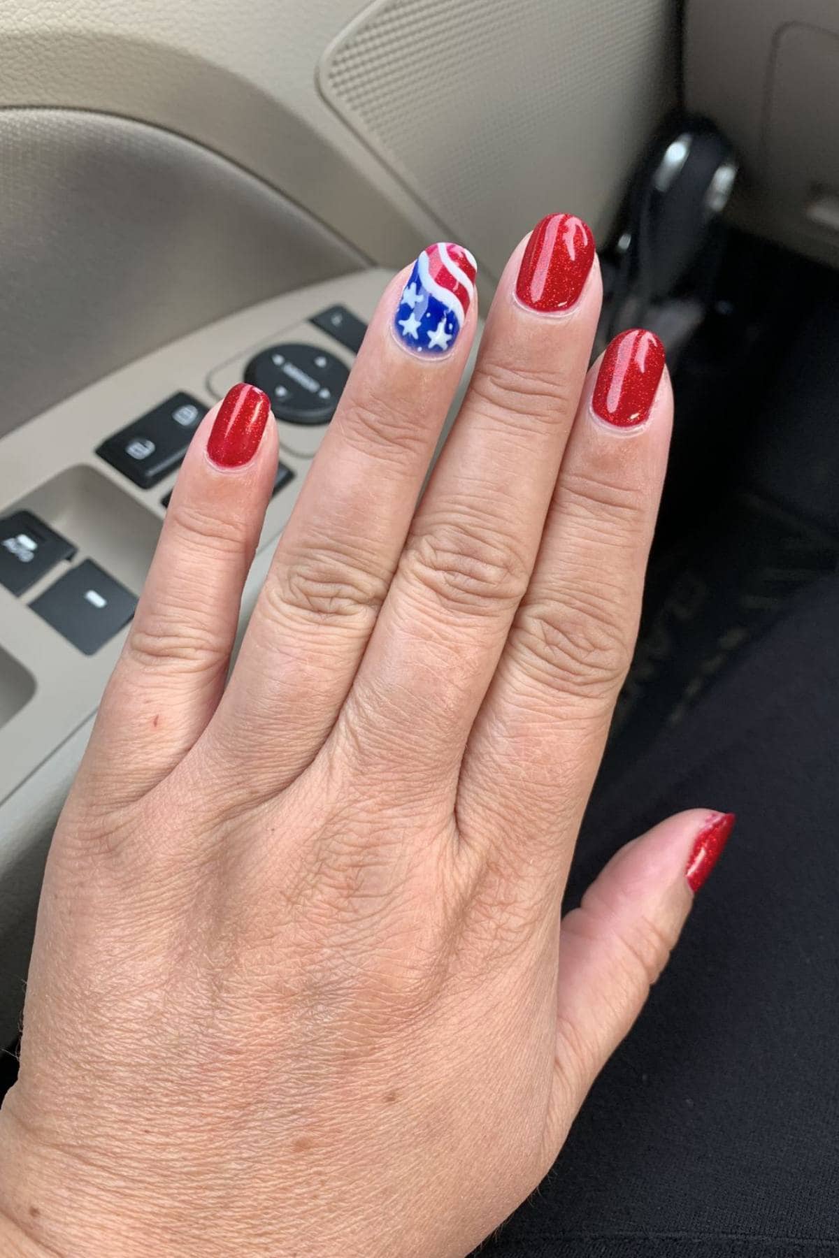 A hand with red glitter nail polish on four fingers and an American flag design with stars and stripes on the ring finger, shown inside a car.