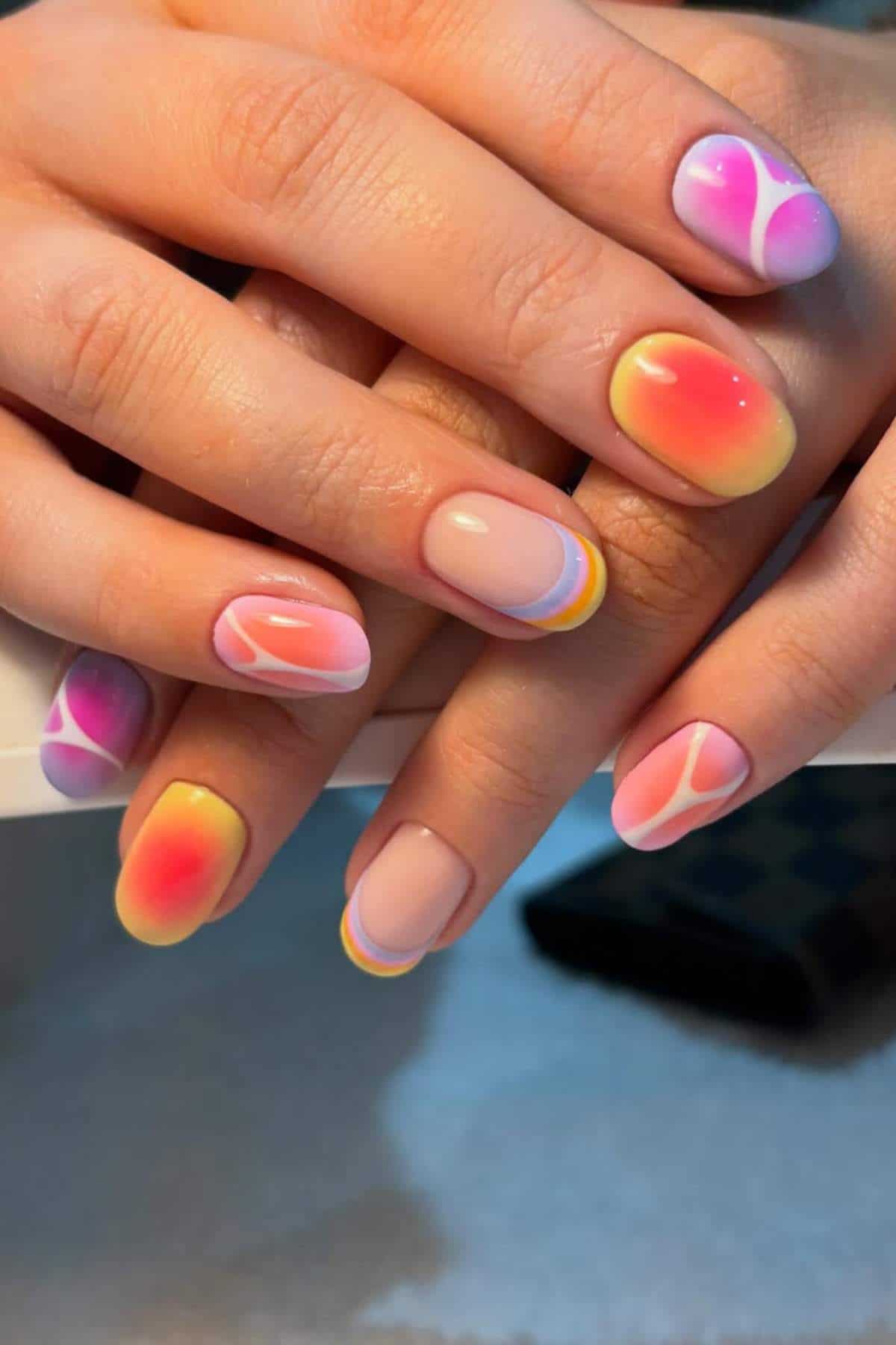 Hands with short nails featuring colorful, abstract swirl and gradient designs in pink, orange, yellow, and blue tones.