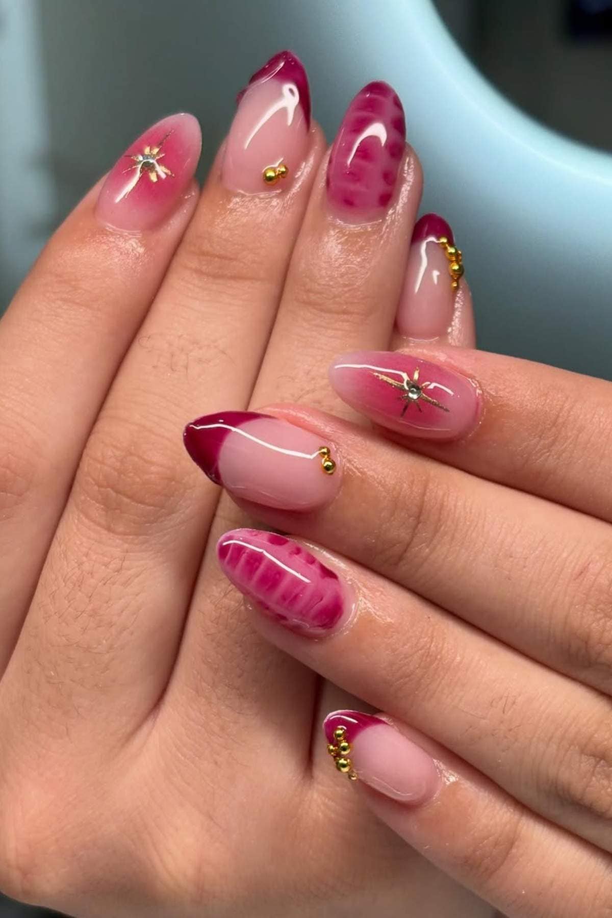 Close-up of hands with long almond-shaped nails featuring pink and burgundy designs, gold bead accents, and small metallic decorations.