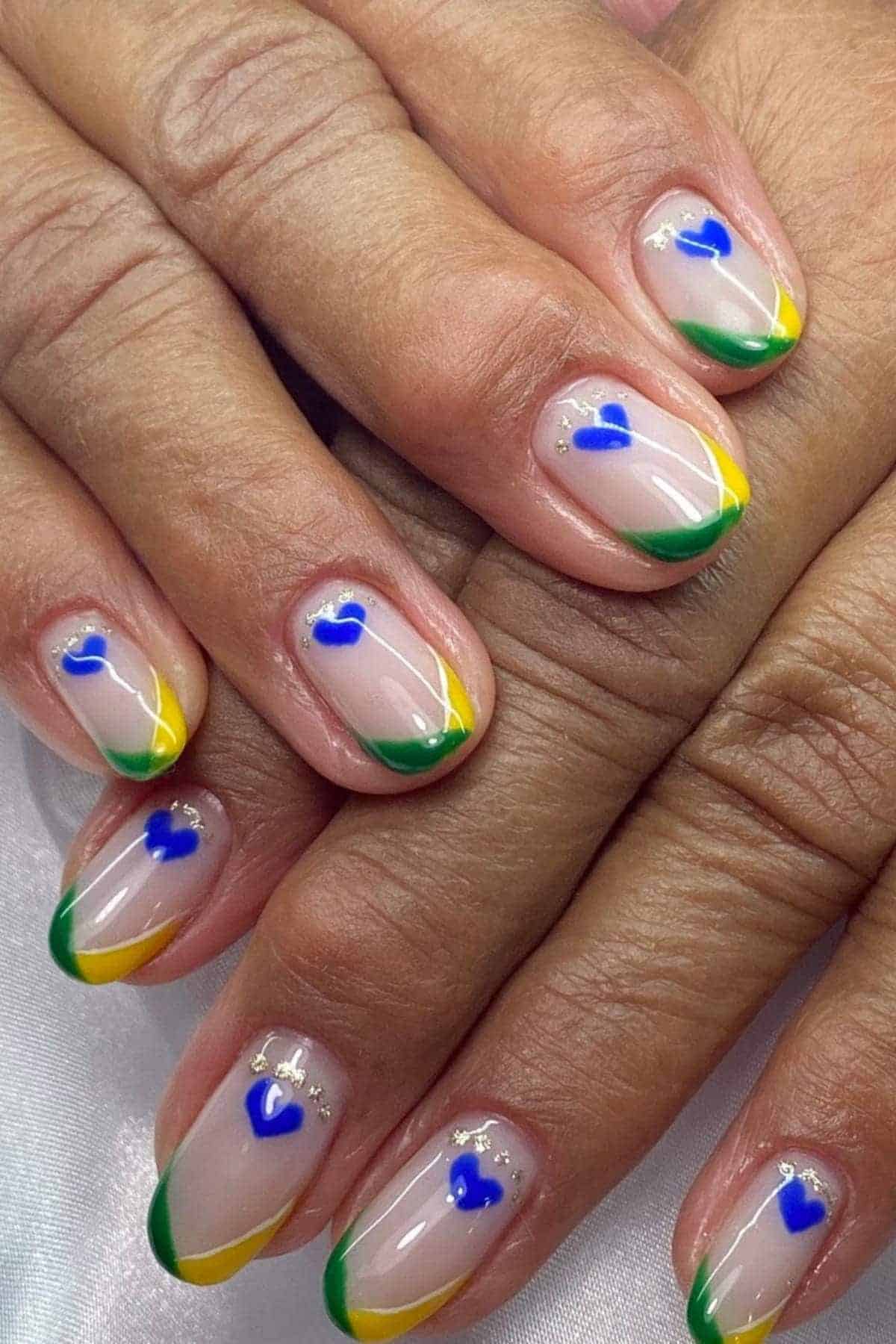 Hands with manicured nails featuring green and yellow French tips and small blue heart designs on a light, natural base.