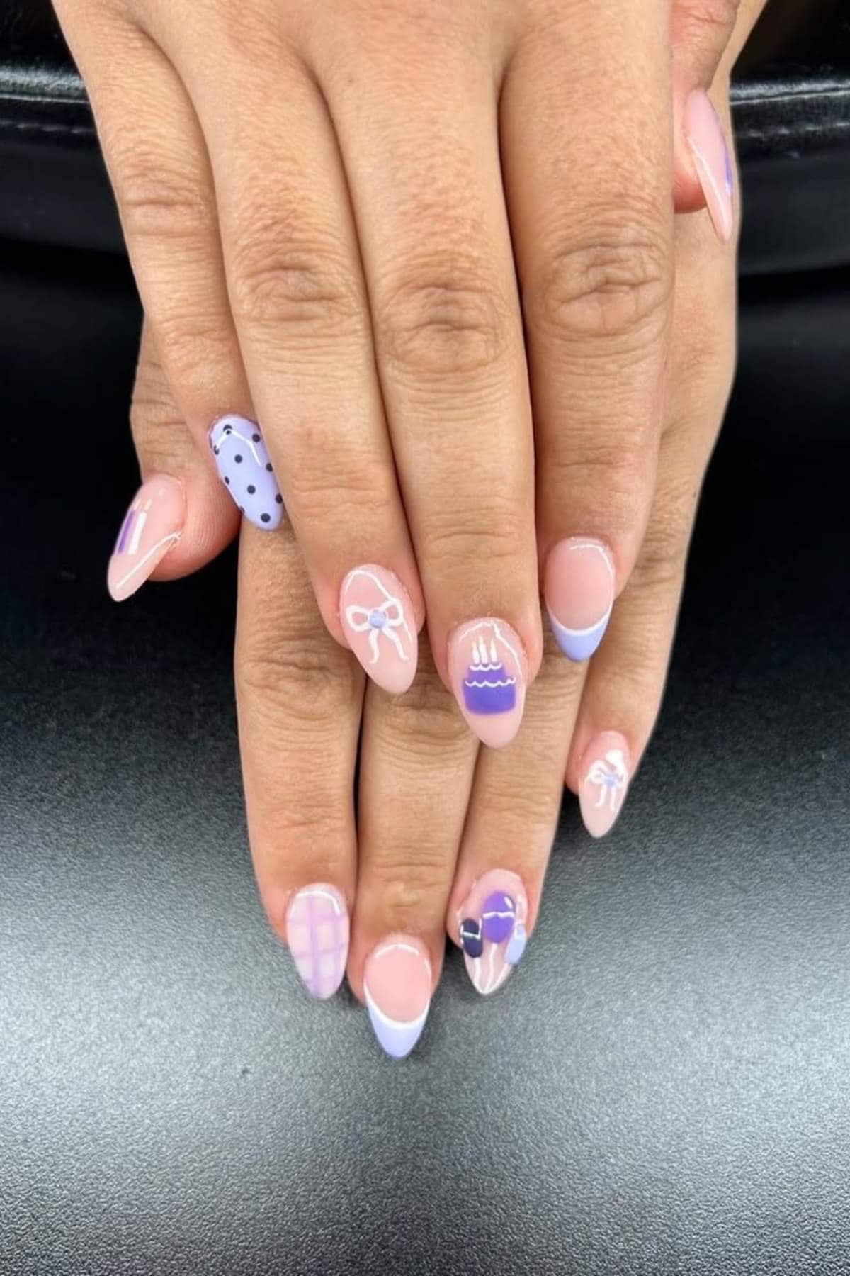 A pair of hands with almond-shaped nails featuring pastel purple nail art, including dots, bows, stripes, and a floral design, resting on a black surface.