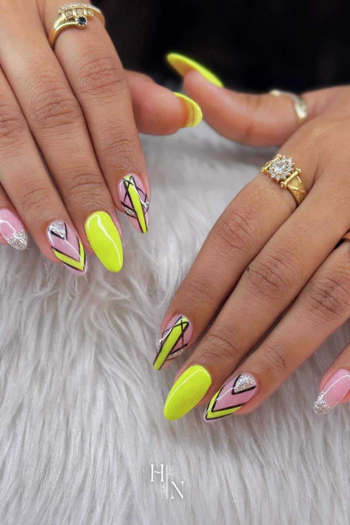 Hands with long, almond-shaped nails featuring neon yellow, black, silver glitter, and pink geometric designs; several gold rings are worn on the fingers.