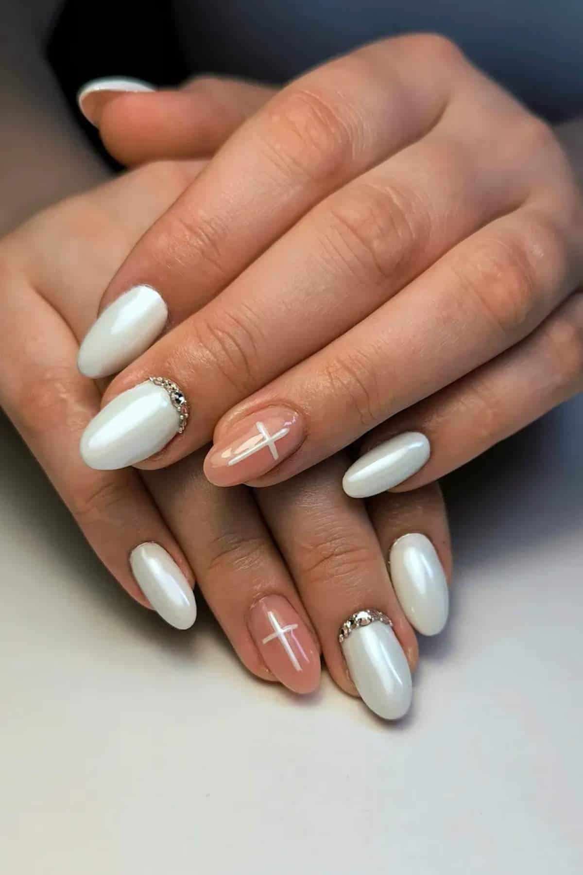 Hands with white almond-shaped nails, accent nails feature a nude base with a white cross, and two nails have small silver embellishments near the cuticles.