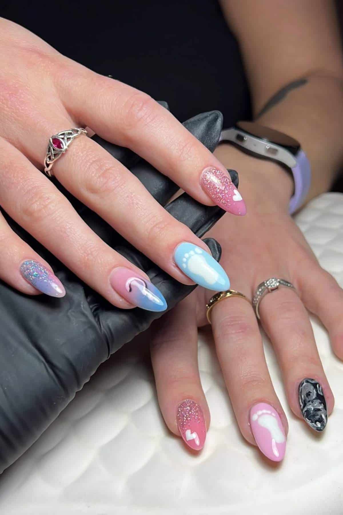 Close-up of hands with long, almond-shaped nails featuring pink, blue, glitter, and black-and-white abstract designs. The person is wearing rings and a smartwatch.