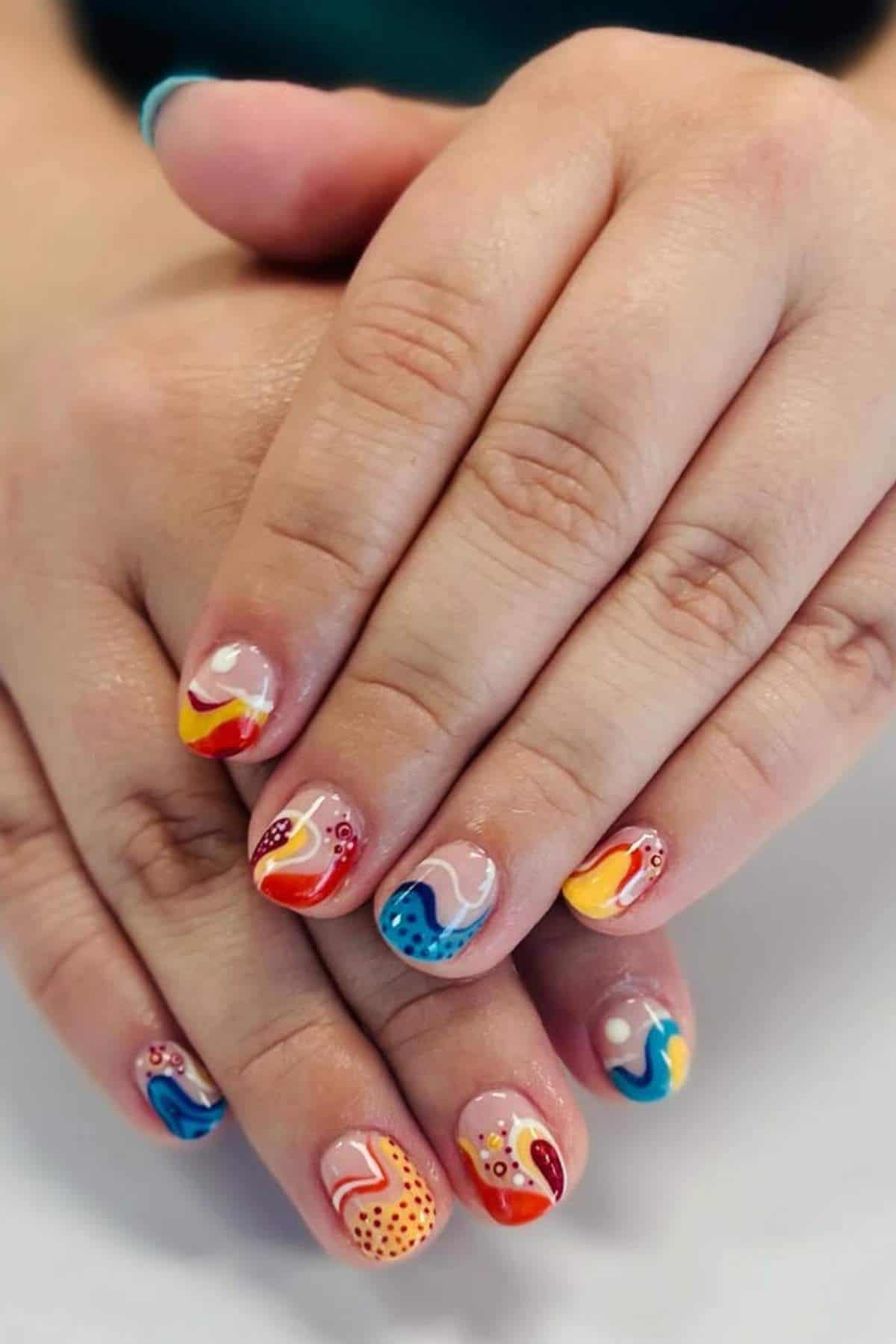 Close-up of hands with short nails painted in colorful abstract patterns, including swirls, dots, and wavy lines in red, yellow, blue, orange, and white.