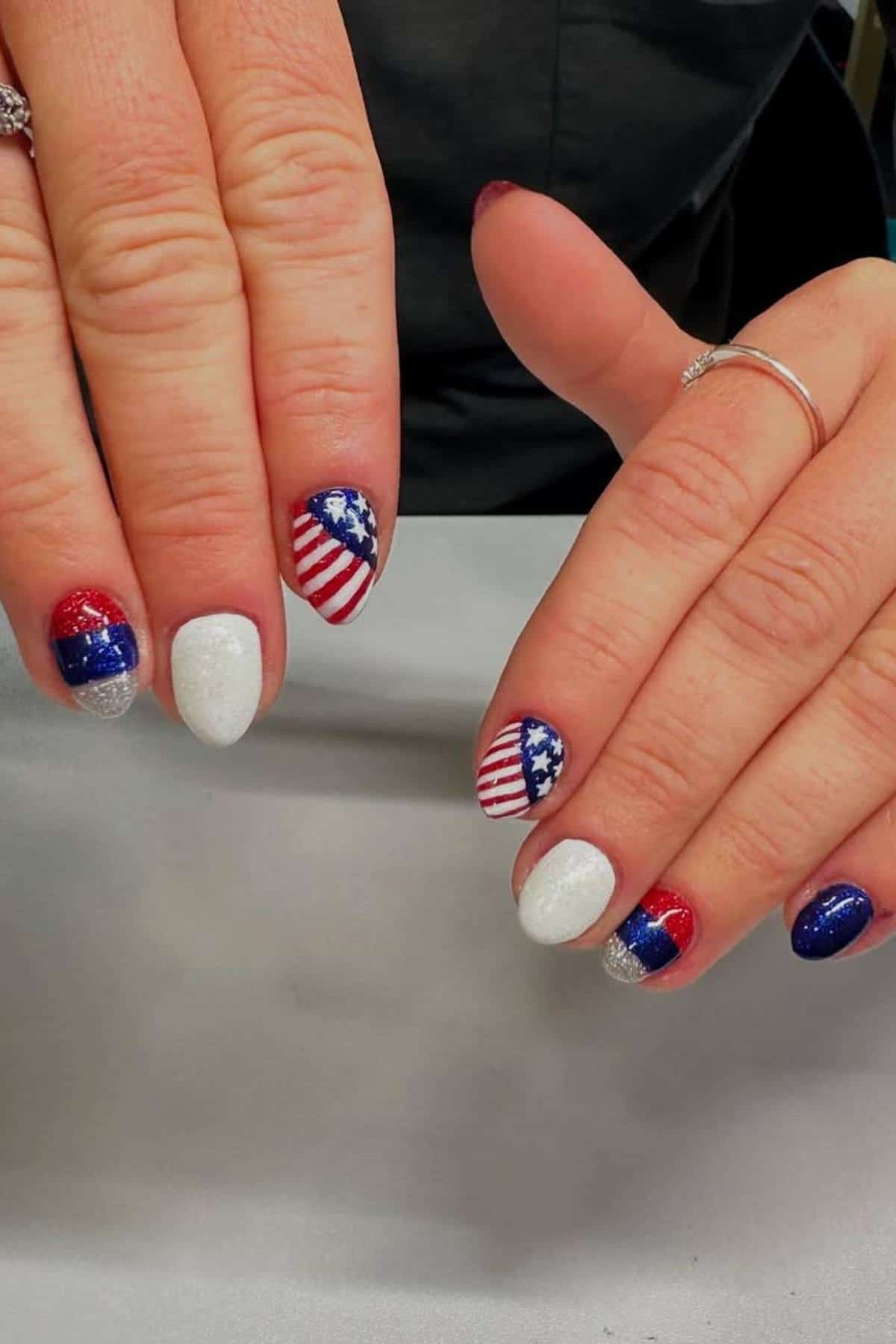 Hands with manicured nails painted in red, white, and blue with glitter and American flag designs, resting on a gray surface.