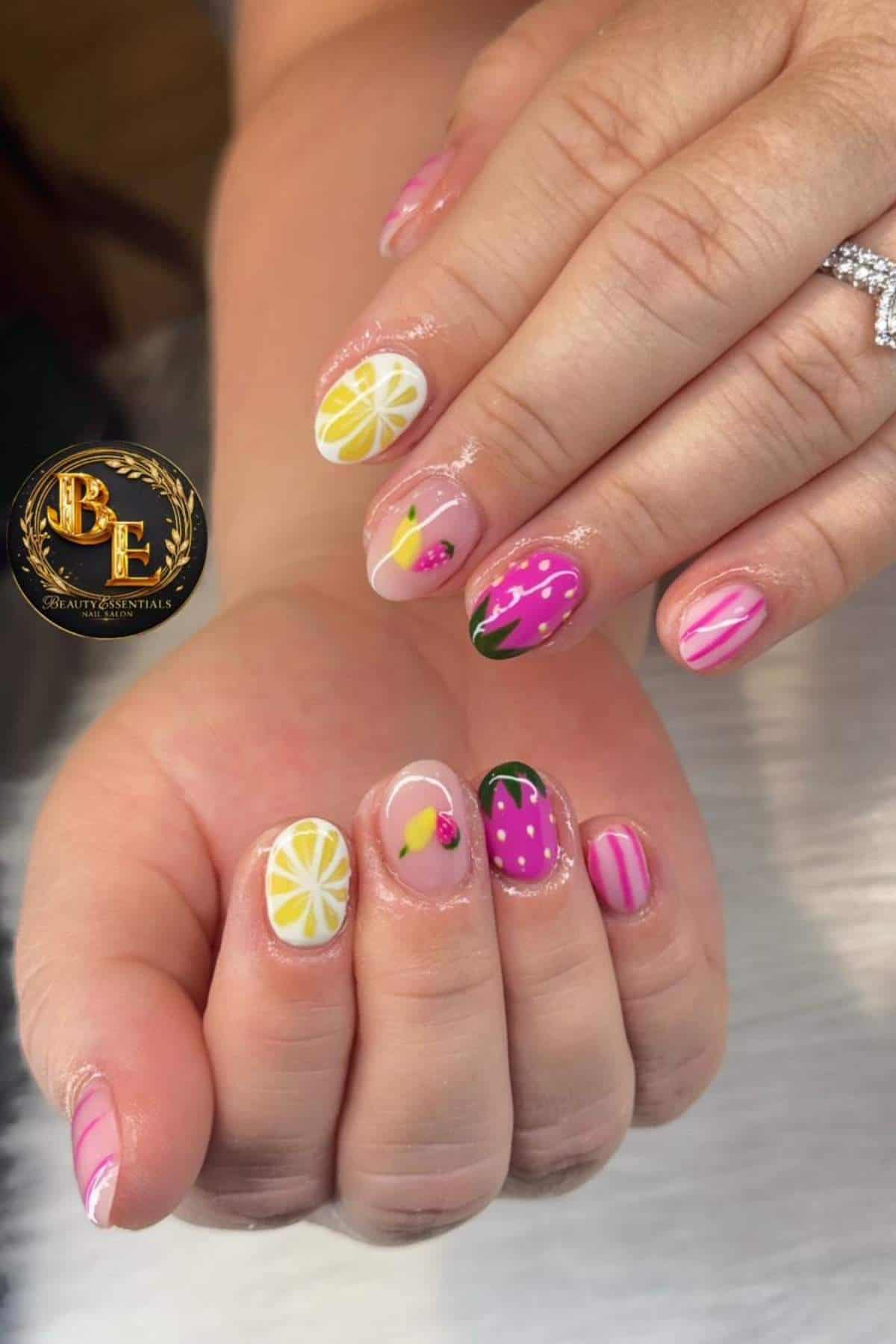 Hands with colorful nail art featuring fruit designs such as lemon, strawberry, cherry, and pink stripes; person wearing a diamond ring.