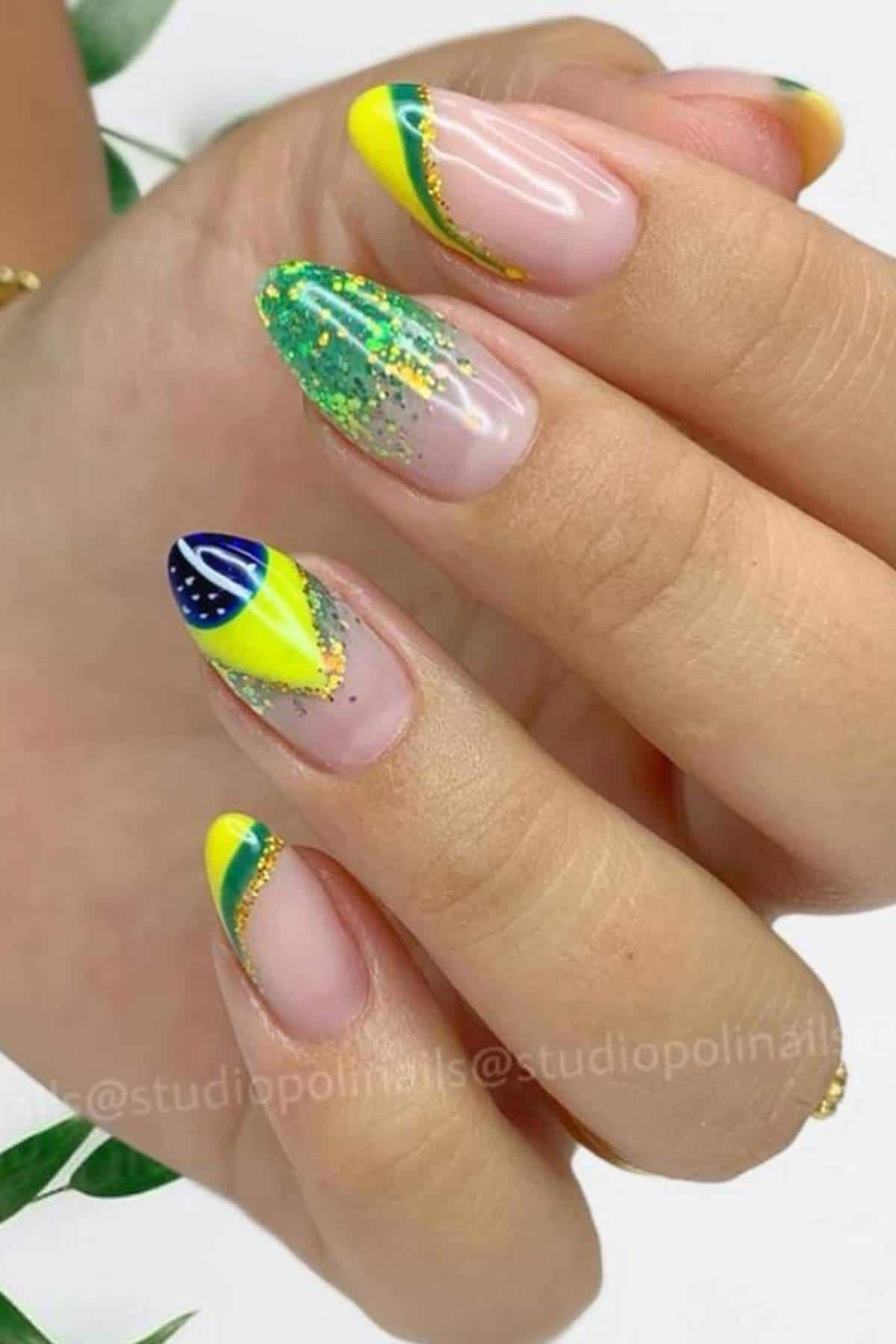 A hand with almond-shaped nails painted with yellow, green, and blue designs, including glitter and a pattern resembling the Brazilian flag.