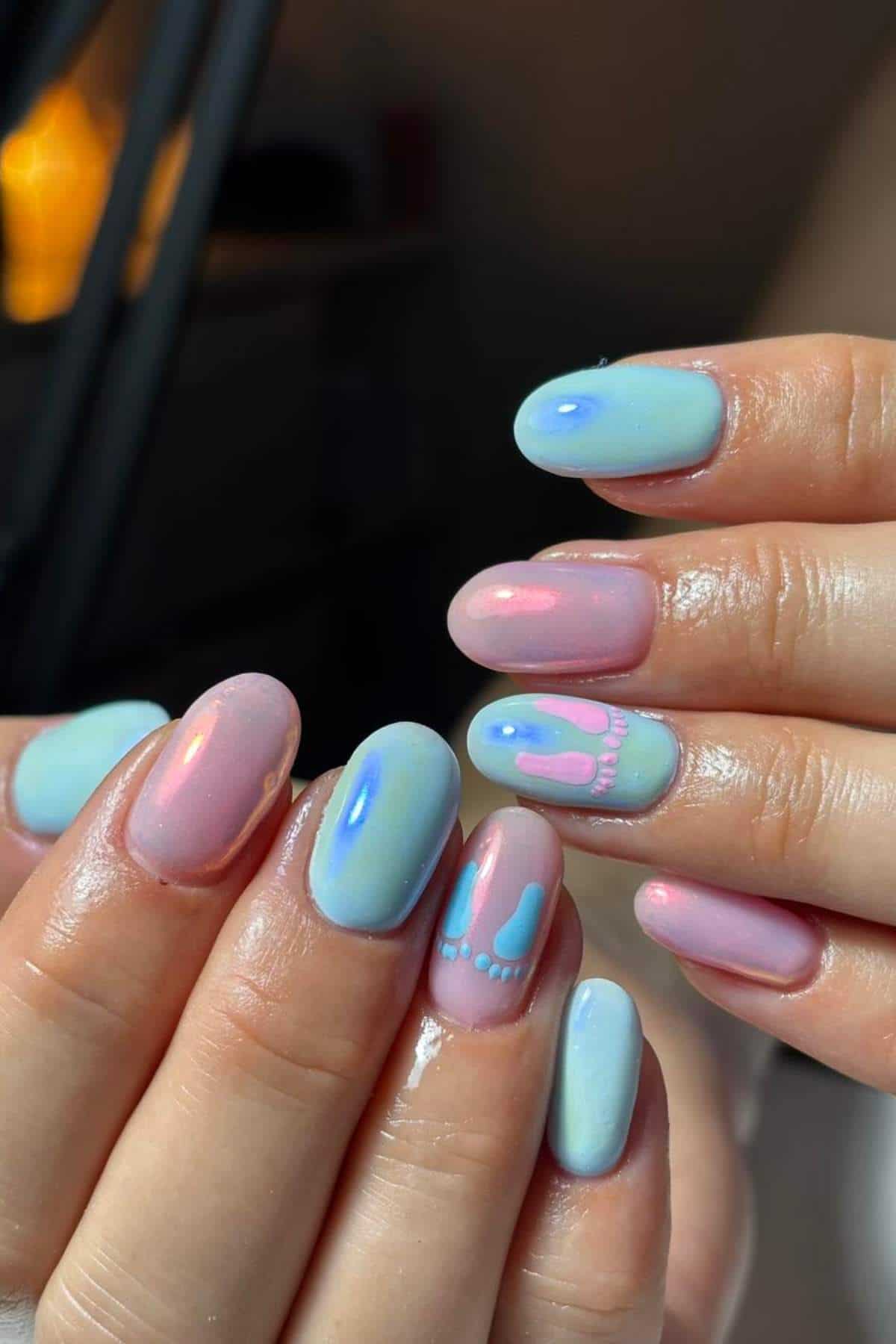 Hands with oval-shaped nails painted in pastel blue and pink, featuring small designs of baby feet and footprints on two accent nails.