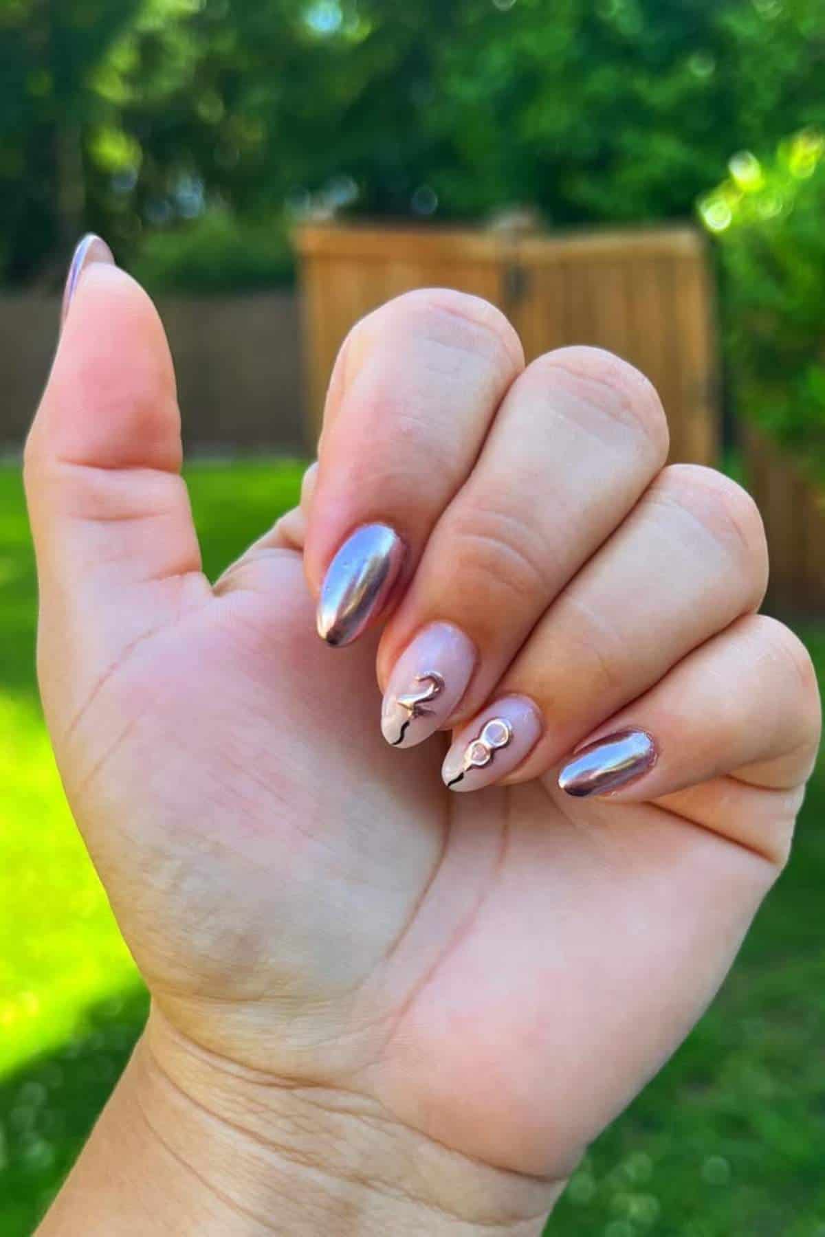 A hand with almond-shaped nails, featuring a shiny chrome finish and two accent nails with floral designs and small gem decorations, is held up outdoors near a wooden fence and greenery.