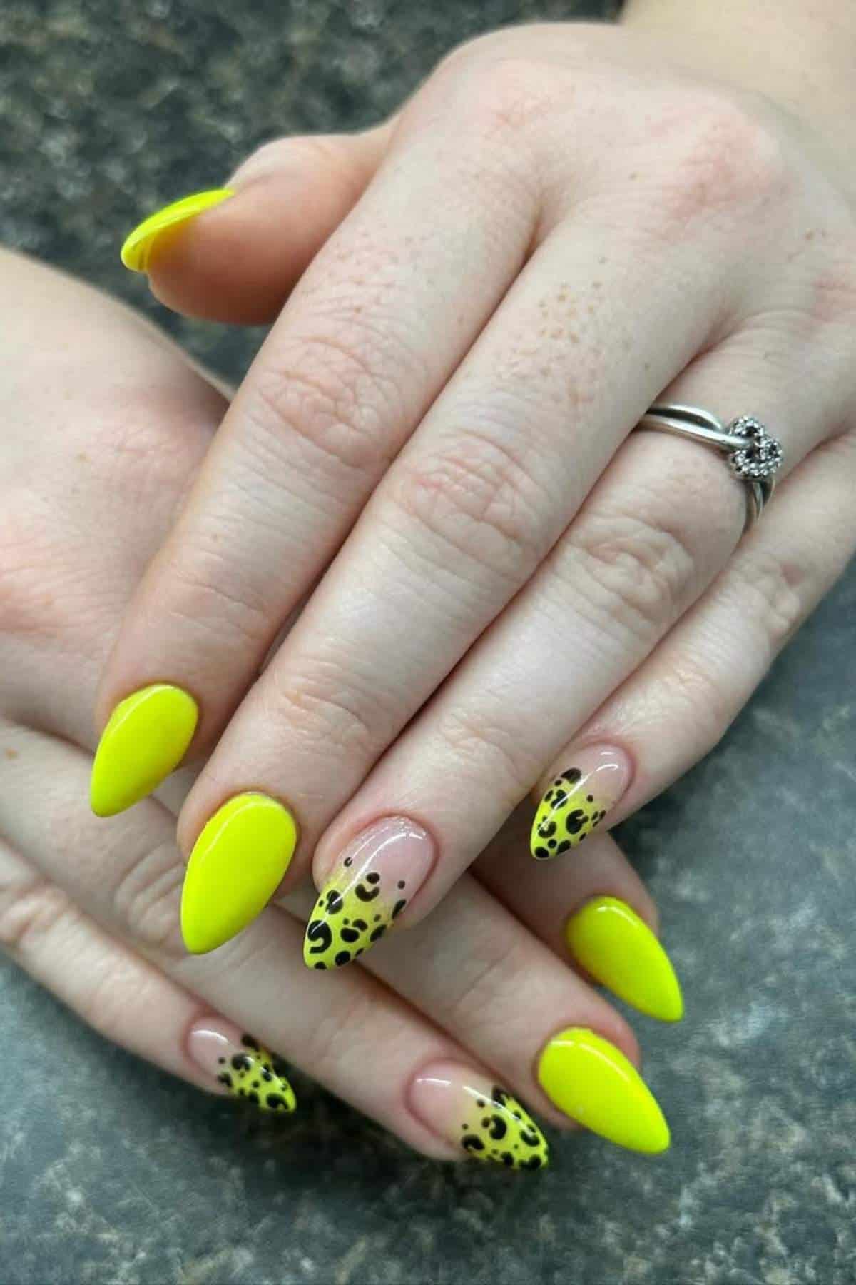 Hands with almond-shaped nails painted bright yellow, with accent nails featuring a yellow and black leopard print design. A ring with a clear stone is worn on the ring finger.