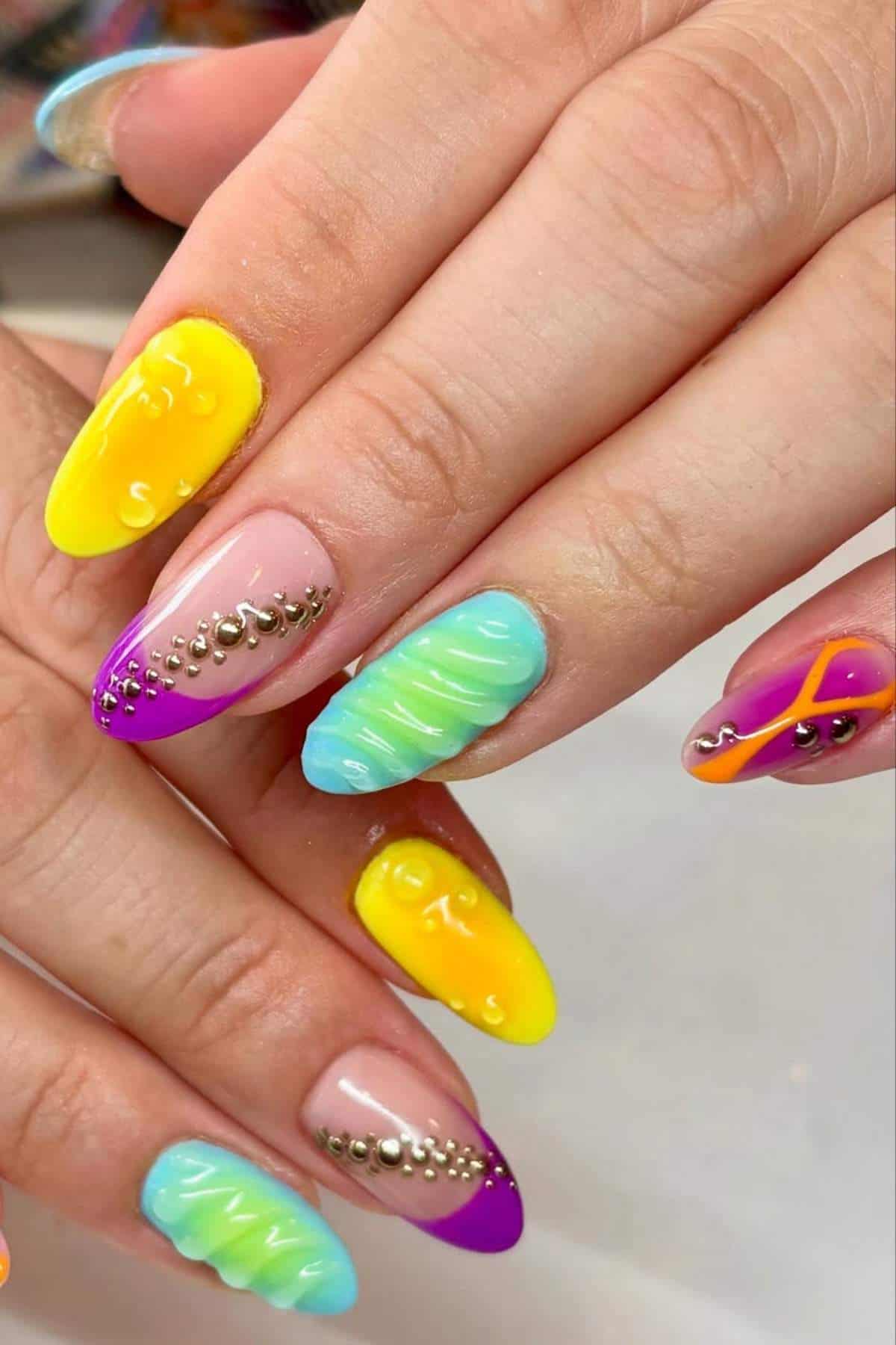 Close-up of hands with colorful, decorated fingernails featuring yellow, blue, purple, and green designs, 3D textures, rhinestones, and geometric patterns.