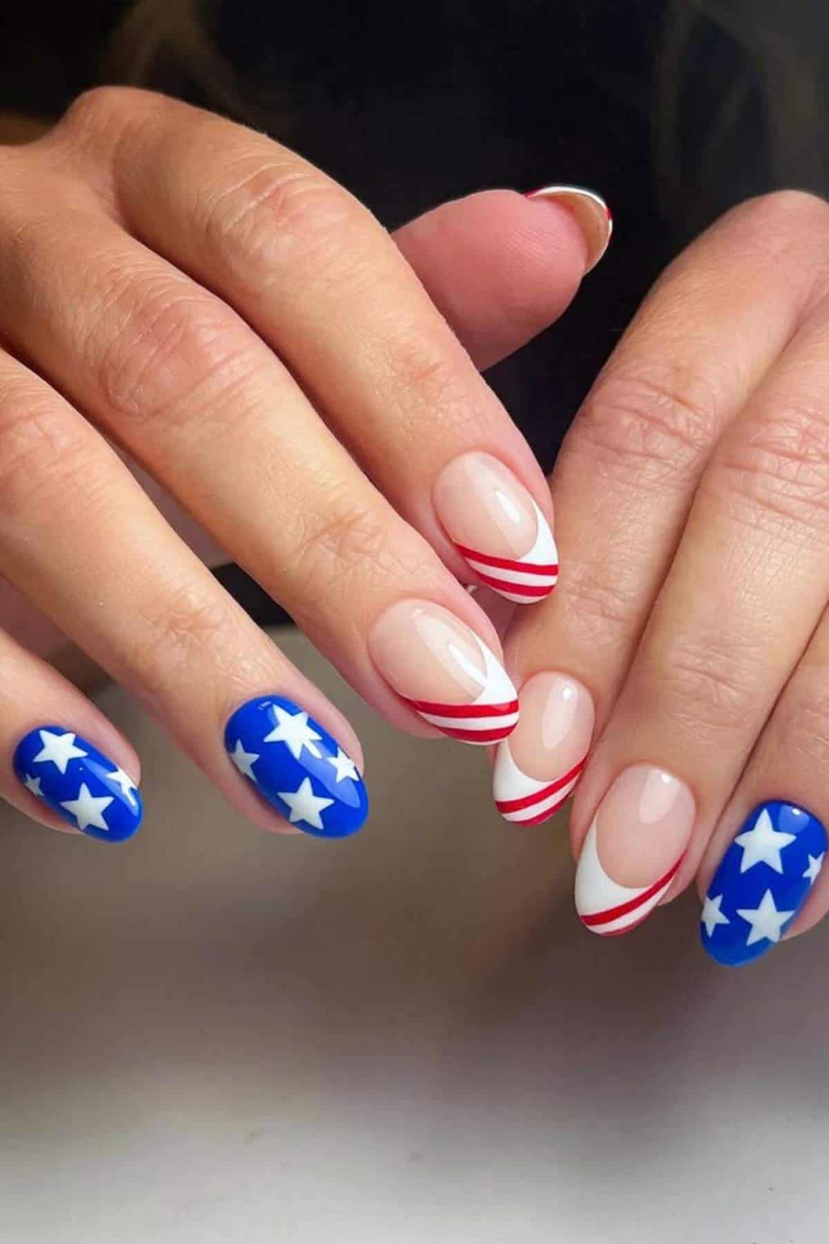 Hands with manicured nails featuring a patriotic design: blue polish with white stars on some nails and red and white striped tips on others, resembling the American flag.