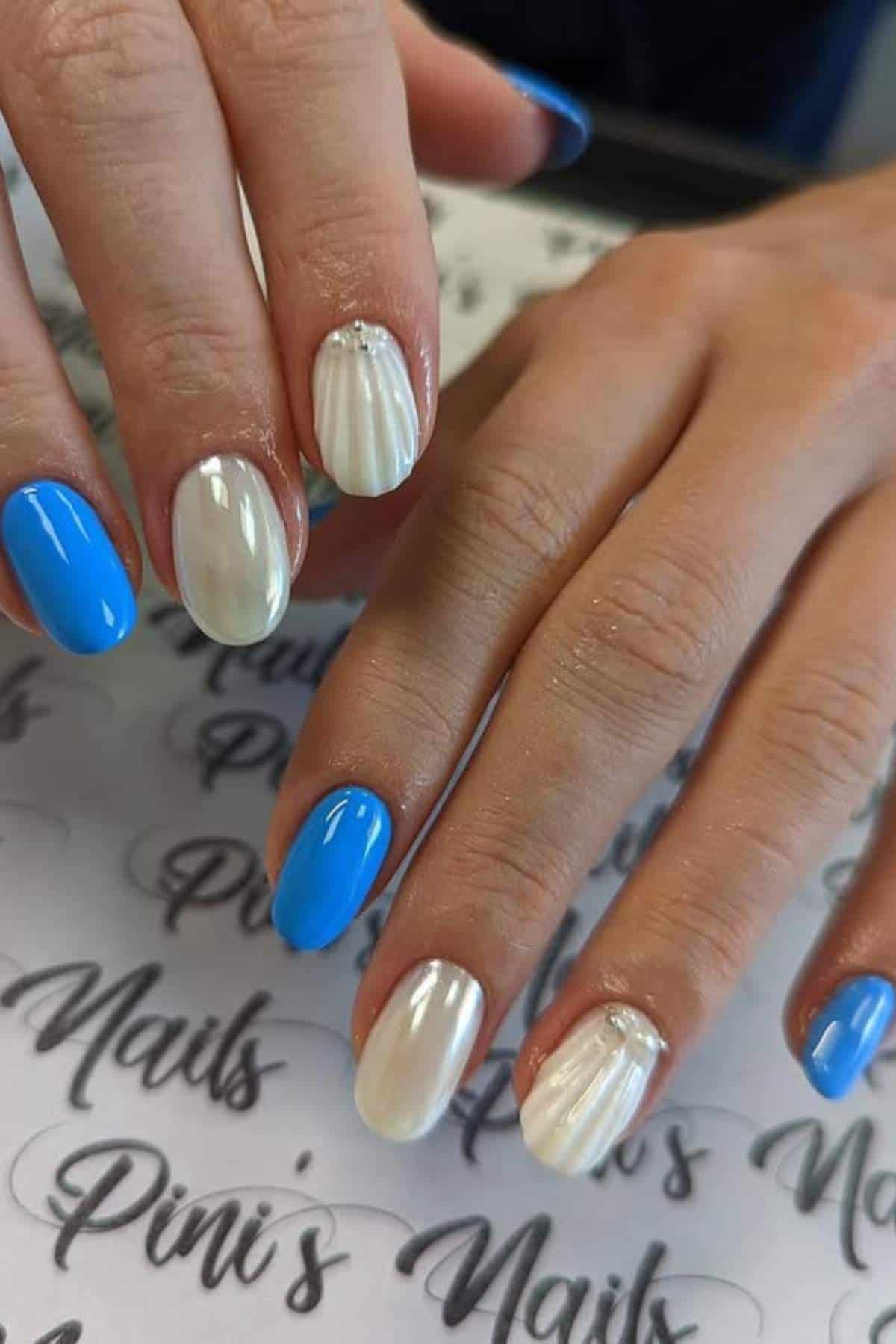 Hands with manicured nails, featuring alternating bright blue and pearly white polish; some white nails have a shell-like textured design.