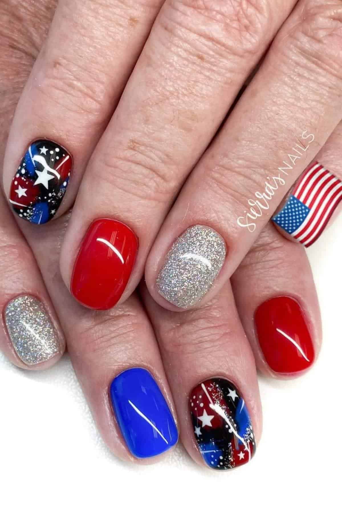 A hand with manicured nails featuring red, blue, silver glitter, and patriotic designs with stars, stripes, and an American flag pattern.
