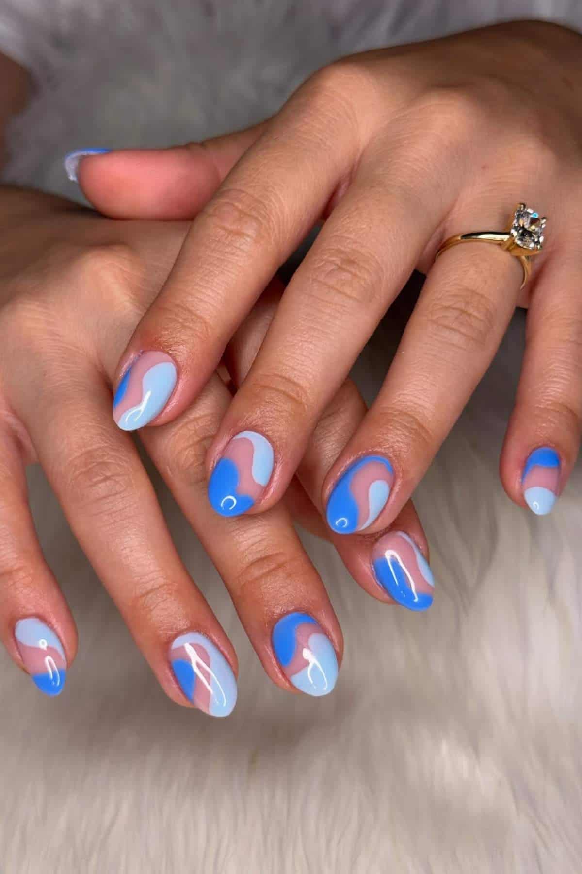Hands with almond-shaped nails painted in blue and light blue wavy designs, resting on a white textured surface. One hand wears a gold ring with a round diamond.