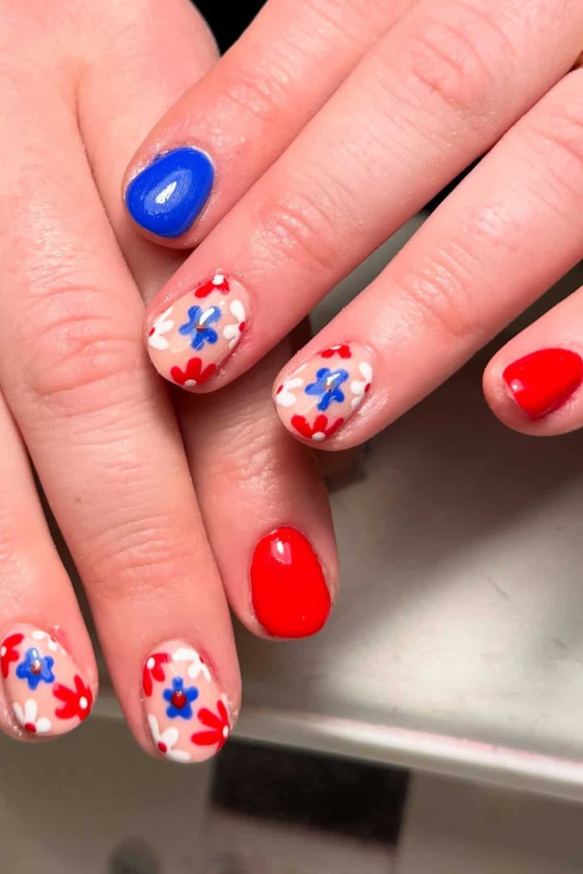 Hands with manicured nails painted in solid blue, solid red, and floral designs in red, blue, and white on a light background.