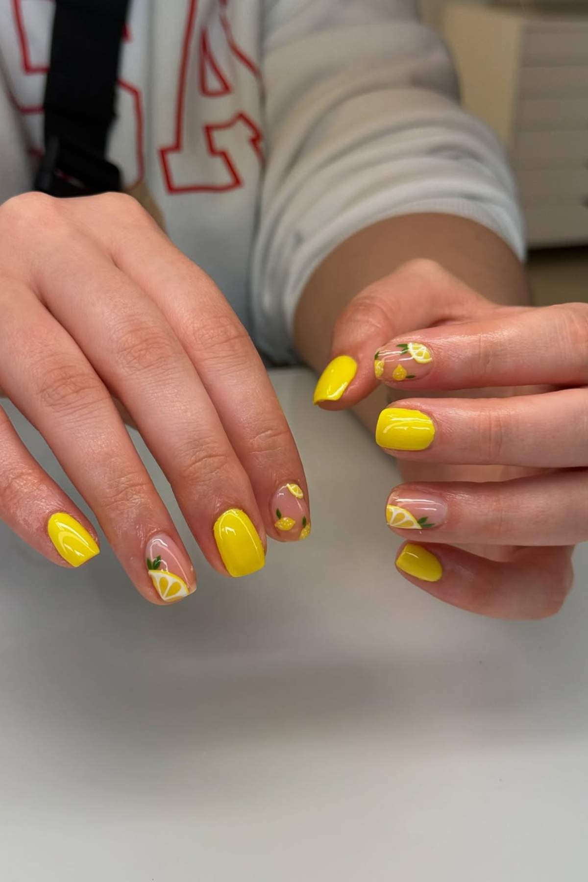 Hands with manicured nails painted yellow, some featuring lemon slice and leaf designs on a few nails, resting on a white surface.