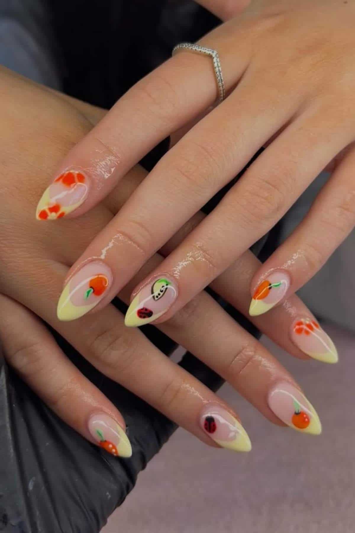 A hand with long, almond-shaped nails featuring yellow tips and small, colorful fruit designs on each nail.