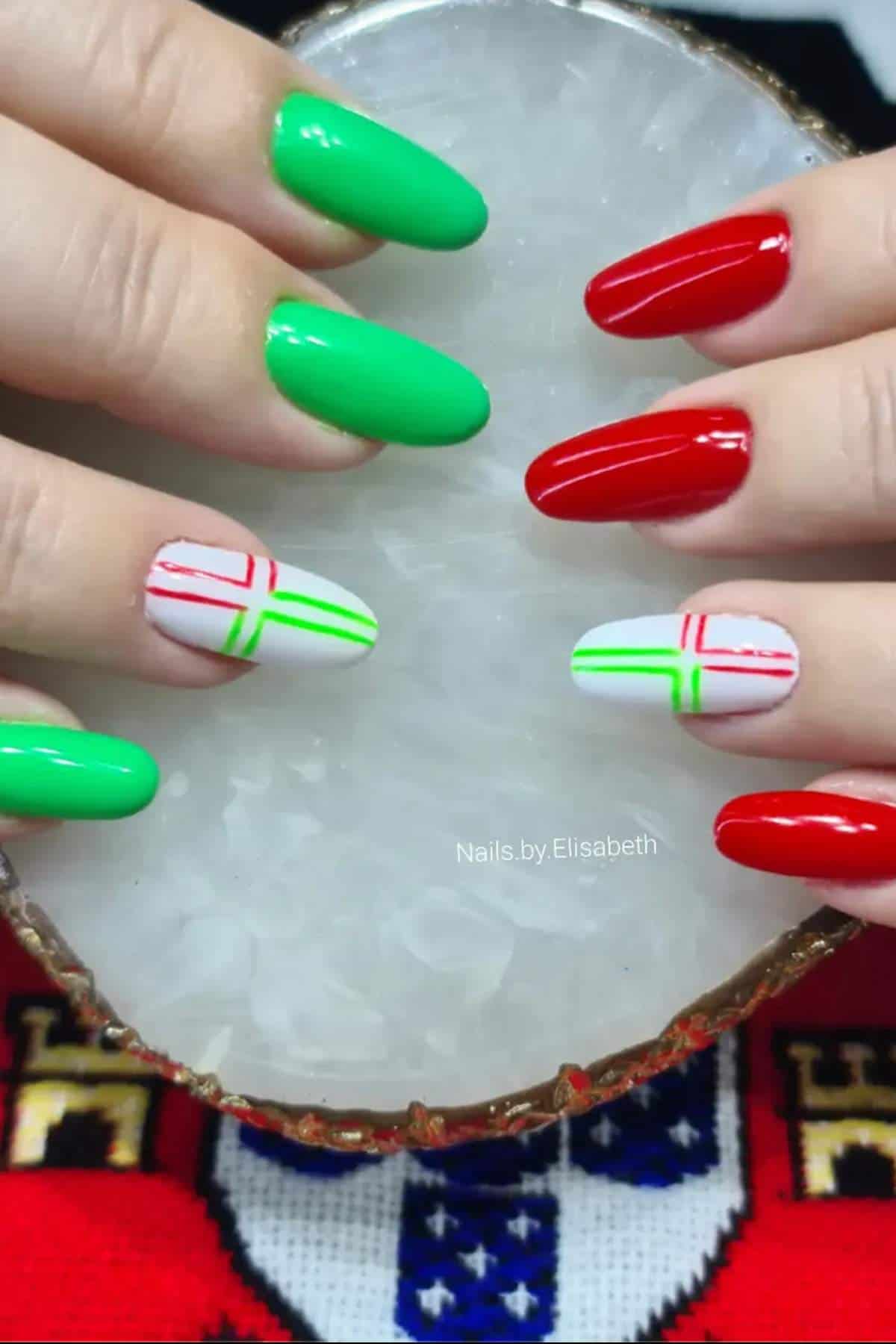 Hands with manicured nails; some nails are painted green, some red, and some white with red and green line designs, displayed against a round white stone.