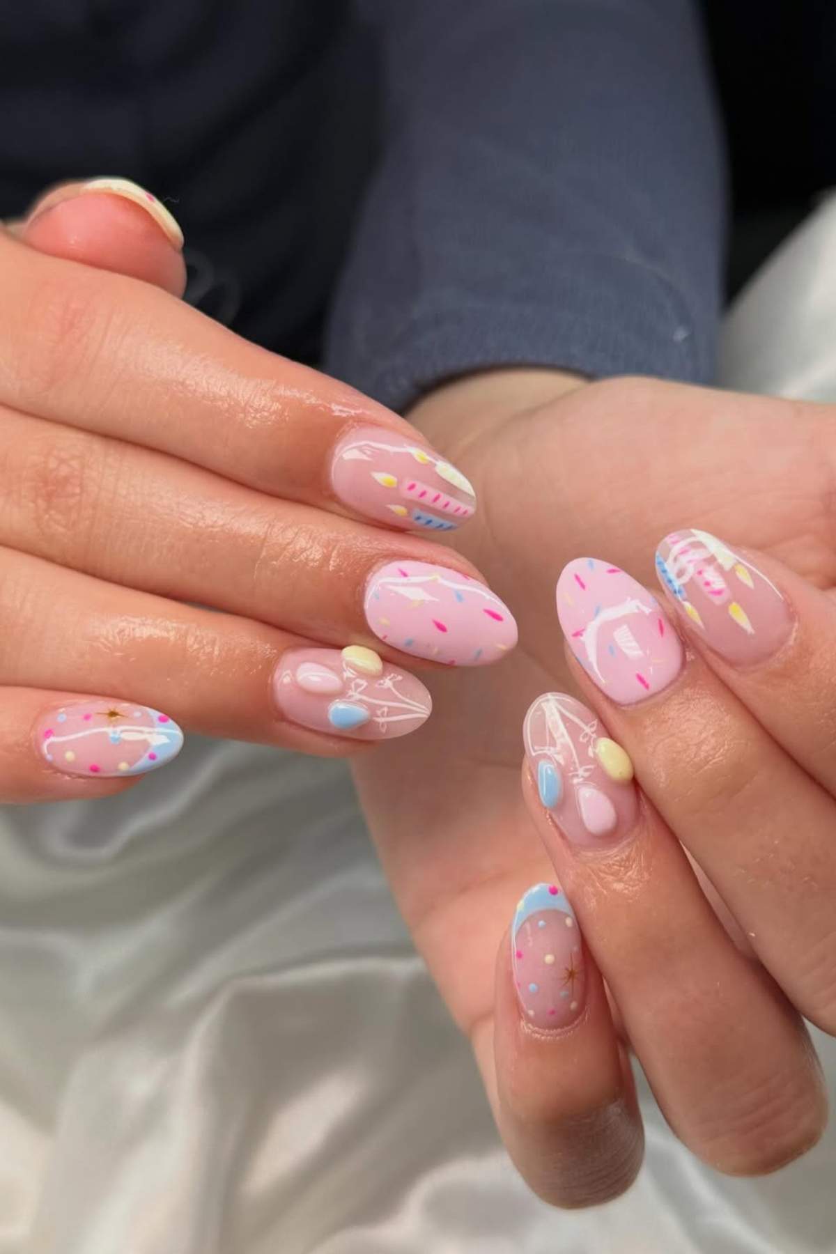 Hands with pastel-colored, almond-shaped nails featuring abstract designs and confetti-like patterns on a light pink base.