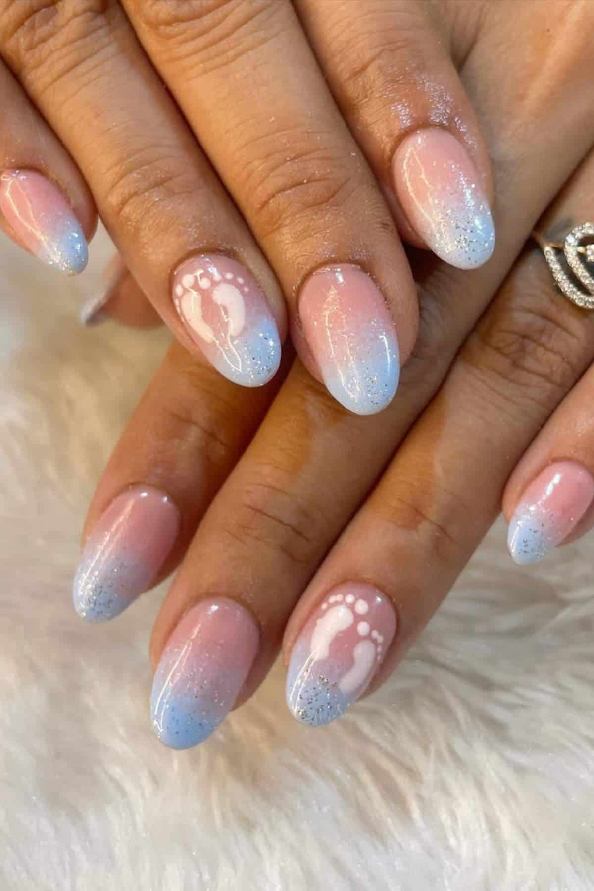 Hands with almond-shaped nails featuring a pink to blue ombre design, glitter accents, and white baby footprint art on two nails.