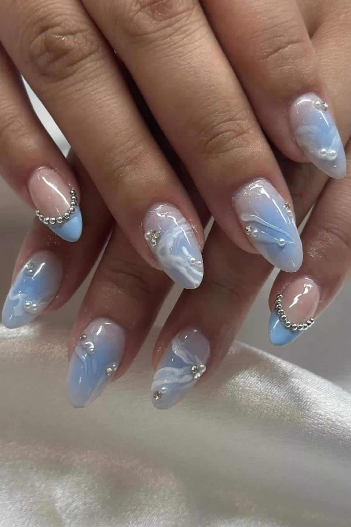 Hands with almond-shaped nails featuring a blue and white marble design, silver bead accents, and glossy finish, resting on a white satin fabric.