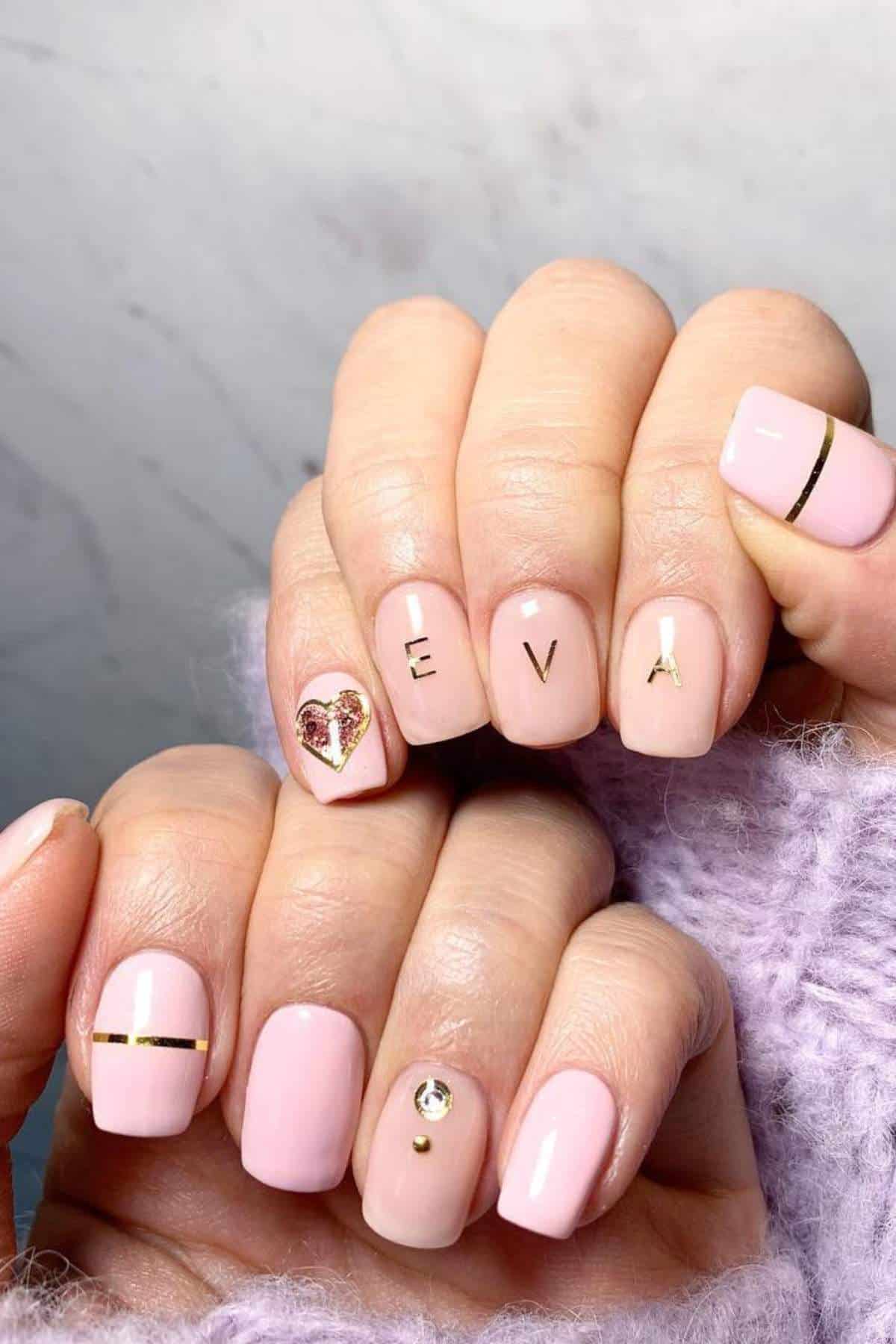 Hands with light pink manicured nails, featuring gold accents, a heart design, and the letters "EVA" on three fingernails.