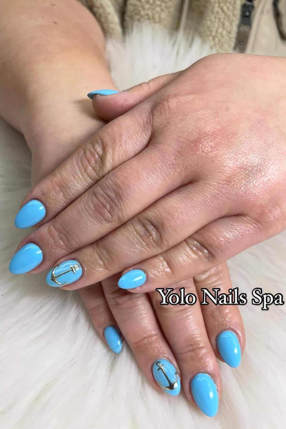 Hands with almond-shaped, glossy light blue nails; two nails feature gold and silver anchor designs. "Yolo Nails Spa" text is overlaid on the image.