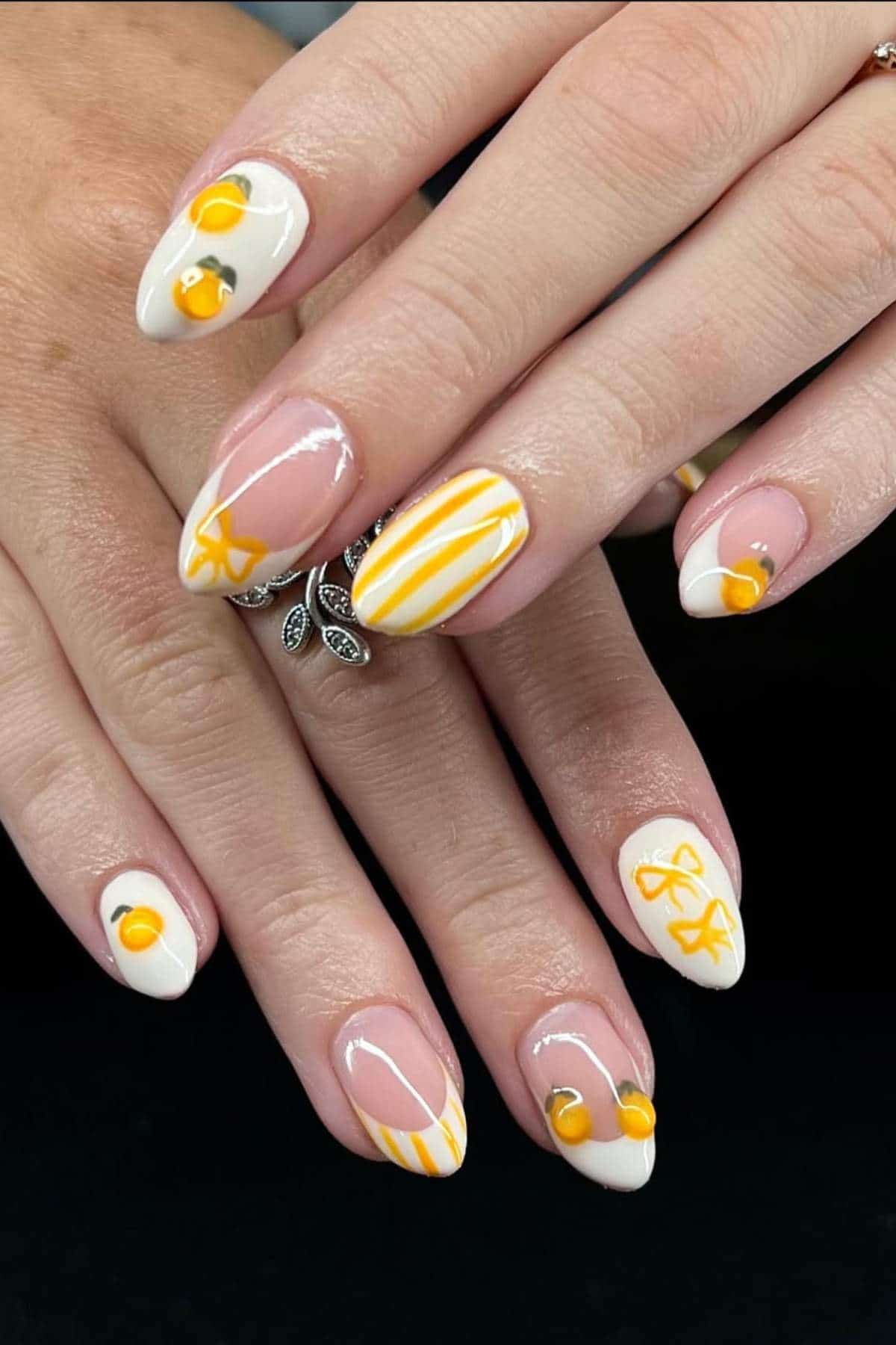 Hands with almond-shaped nails featuring white, yellow, and clear designs, including stripes, dots, and abstract shapes. One hand wears a silver ring with leaf details.