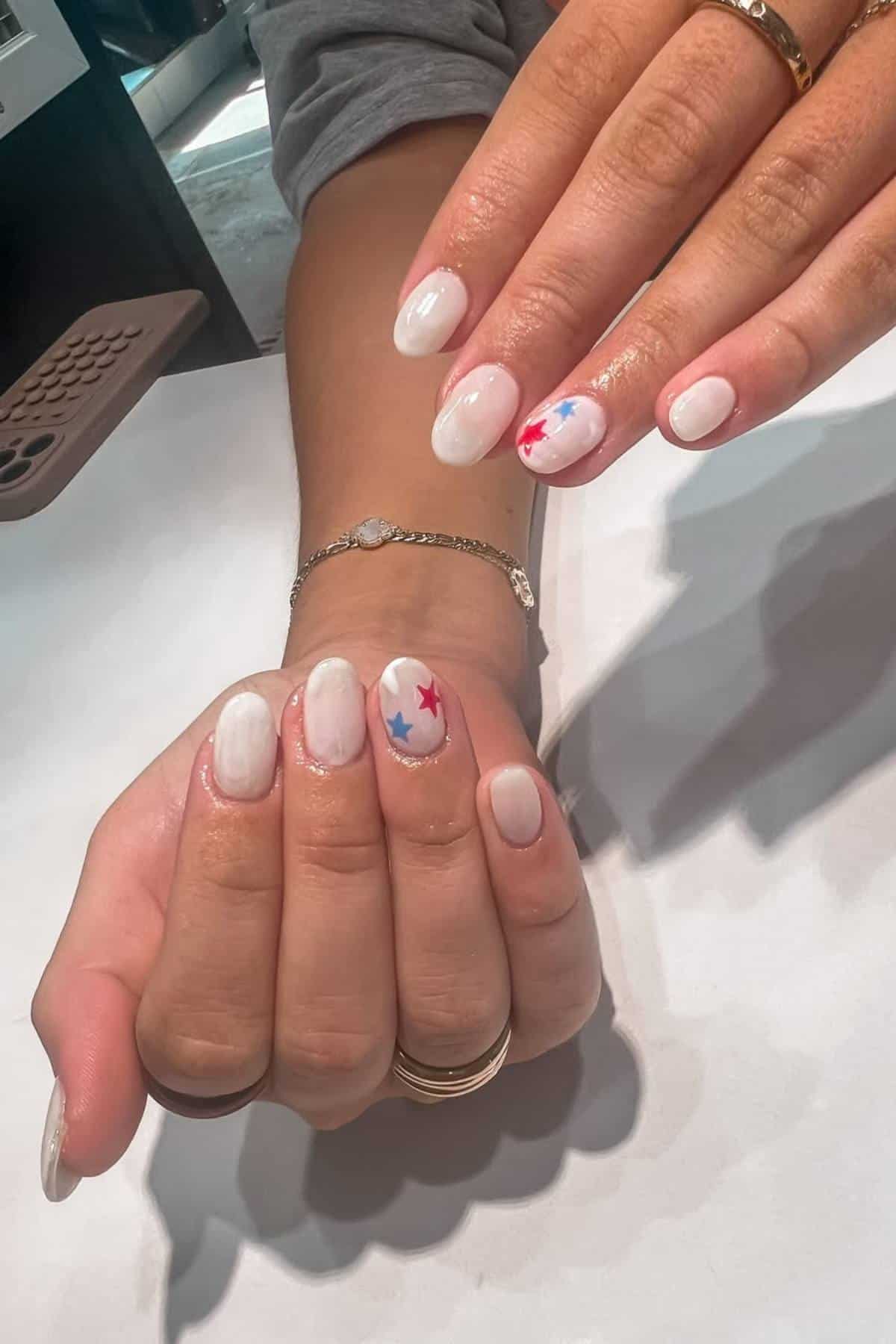 Hands with oval-shaped, glossy white nails; some nails have red and blue star decorations. The person wears multiple rings and a bracelet.