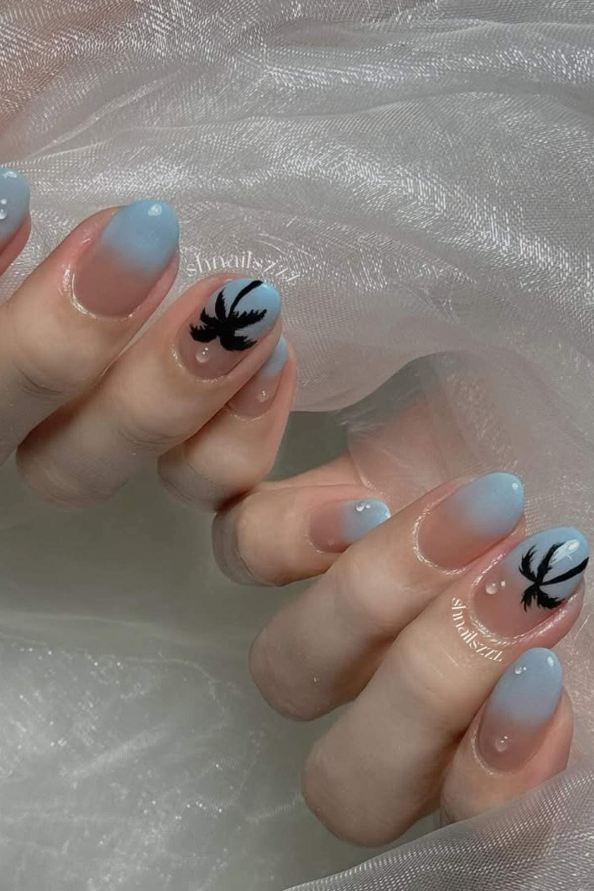 Close-up of hands with almond-shaped nails, painted in a blue gradient with black palm tree designs on two accent nails, resting on a sheer white fabric.