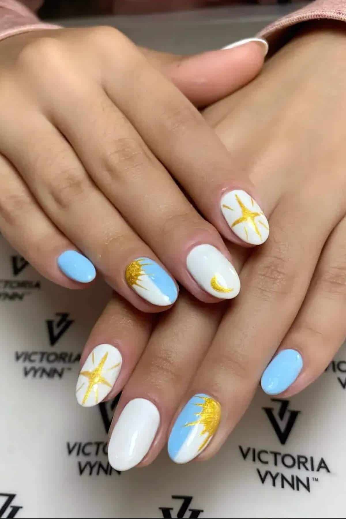 Hands with blue and white manicure, featuring gold sun, star, and crescent moon designs on some nails. Fingernails rest on a Victoria Vynn branded surface.