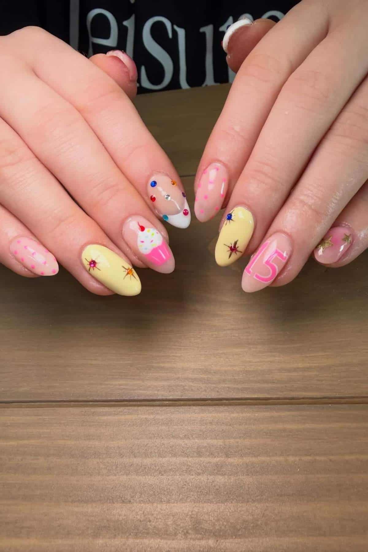 Hands with long almond-shaped nails featuring pastel yellow, pink, and white nail art, including a cupcake, stars, the number 5, and colorful dots, displayed on a wooden surface.