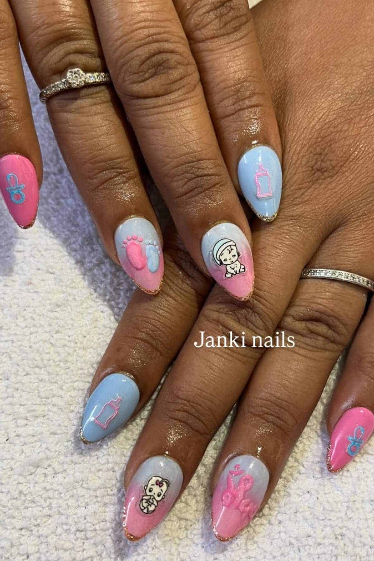 Close-up of hands with blue and pink baby-themed nail art, including bottles, pacifiers, baby faces, and footprints, on almond-shaped nails.