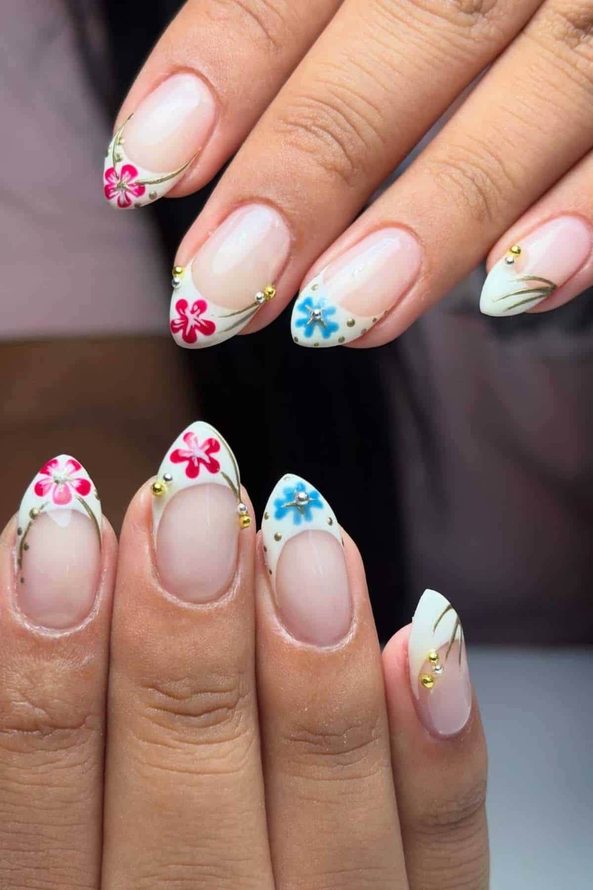 A hand with almond-shaped nails featuring floral nail art in pink and blue, gold accents, and a clear to white gradient base.